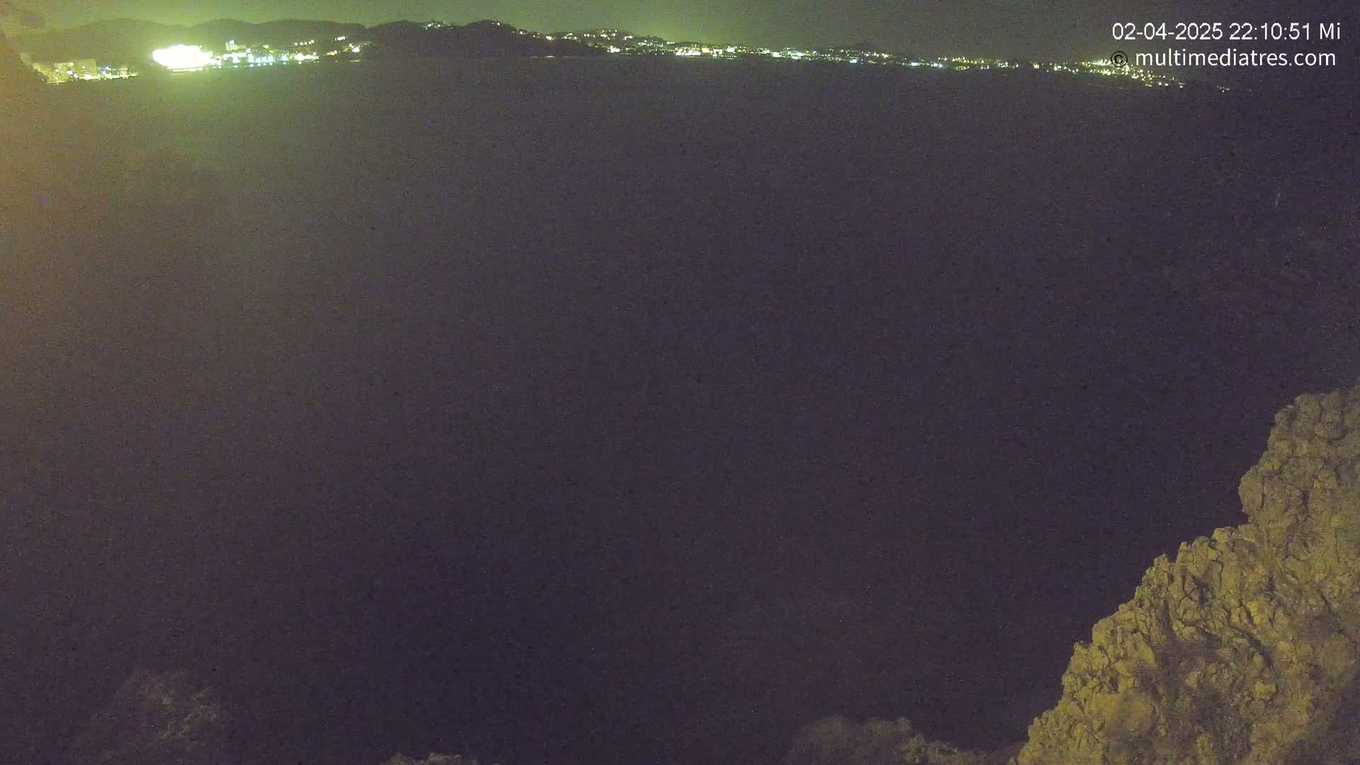 A nighttime view shows a dark body of water with a distant, illuminated cityscape on the horizon and a rocky outcrop in the lower right corner.