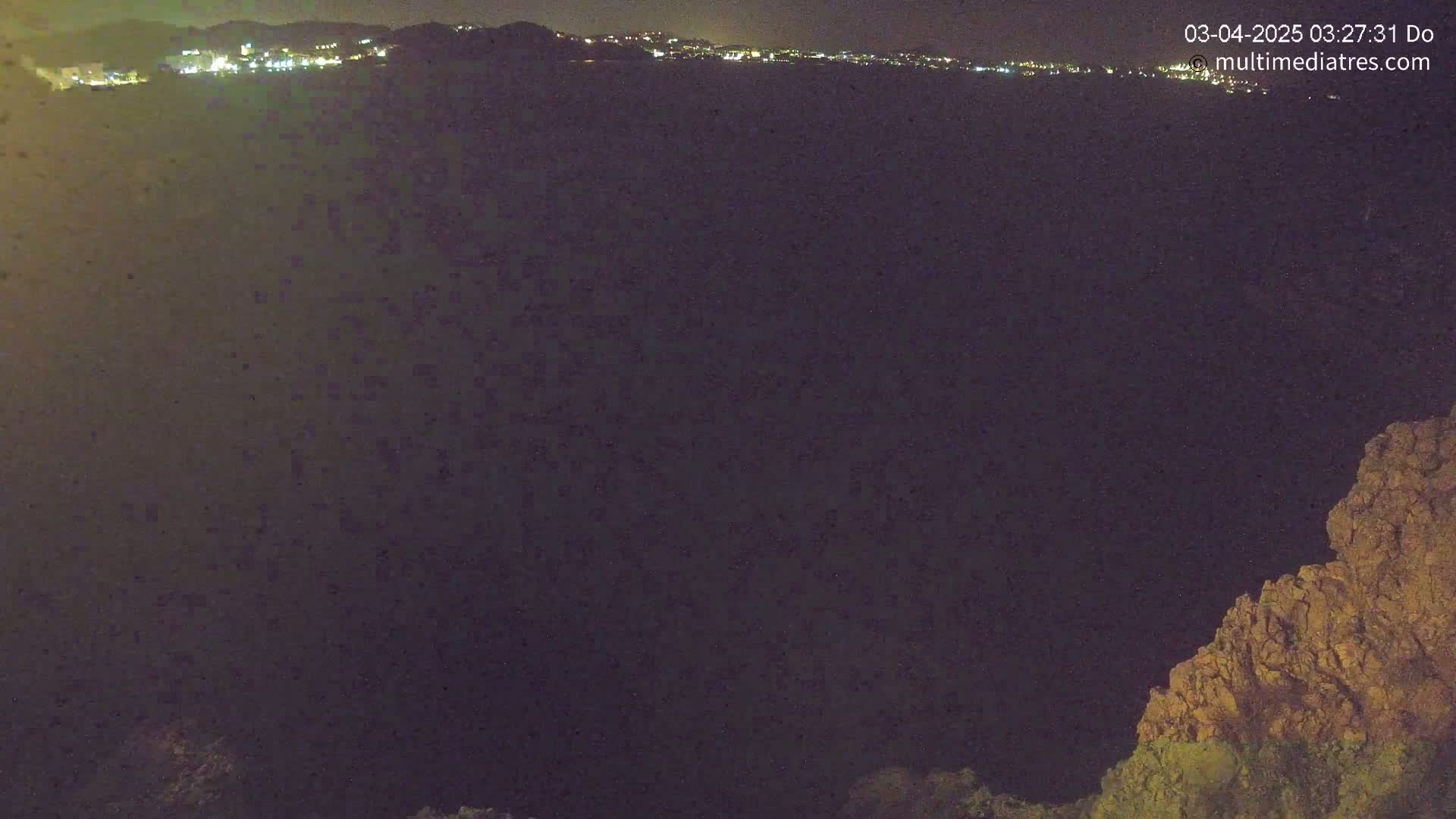 A nighttime, long-range view shows a dark body of water below a distant, faintly lit town, partially framed by a rocky outcrop on the right.