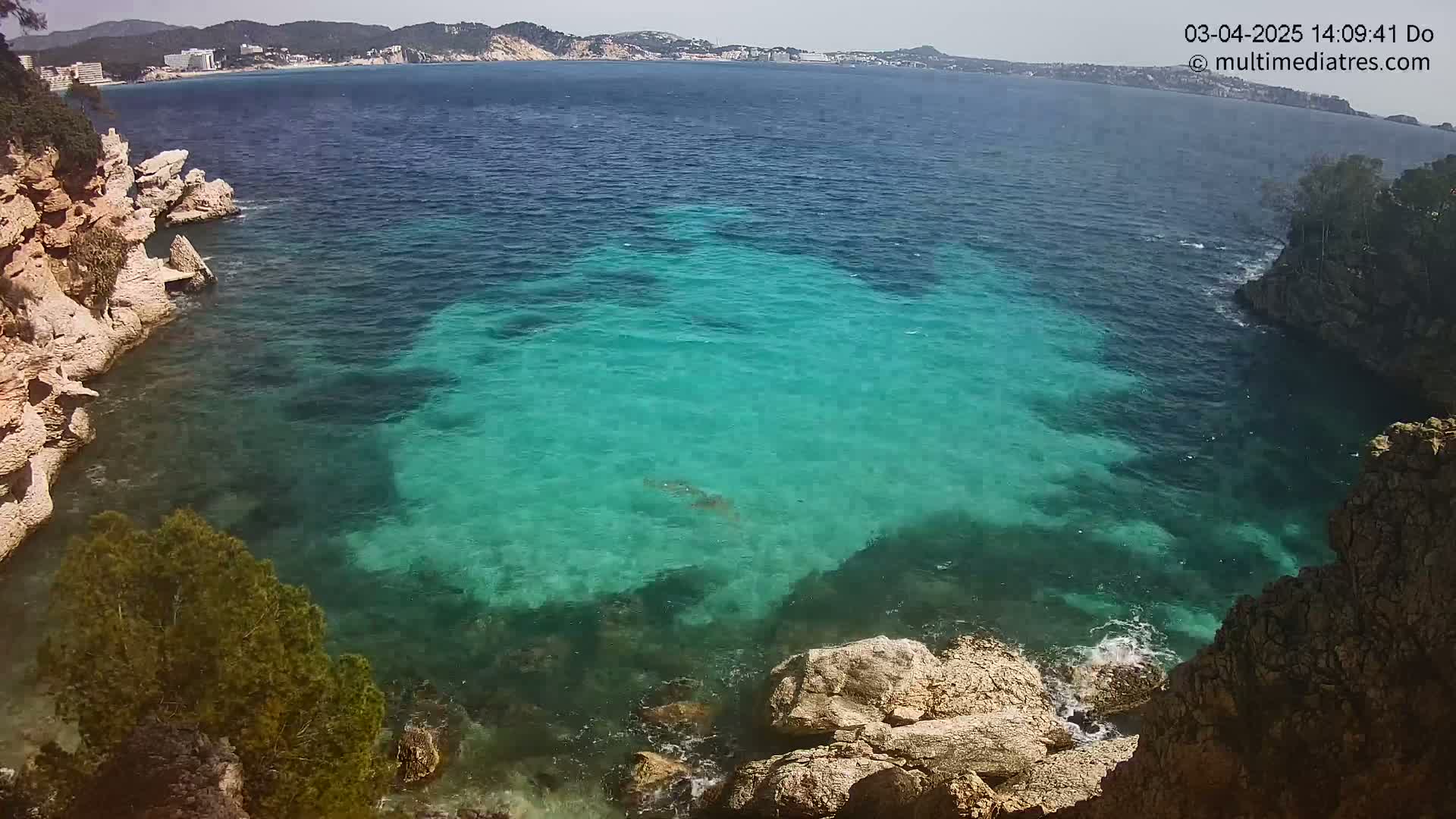 Clear, teal water laps against rocky, light-brown cliffs on a sunny day, with a distant shoreline visible in the background.