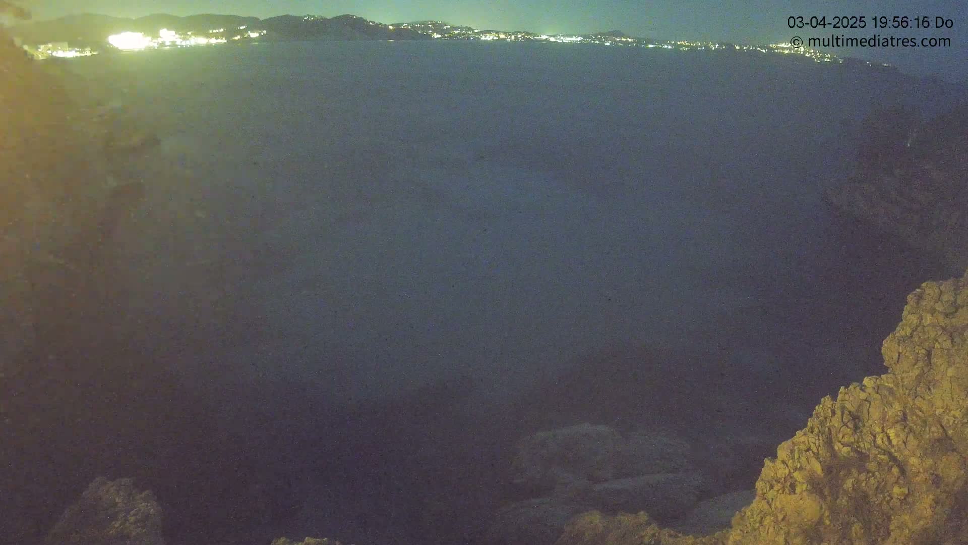 A nighttime view of a dark body of water with a distant city skyline illuminated, observed from a rocky vantage point.
