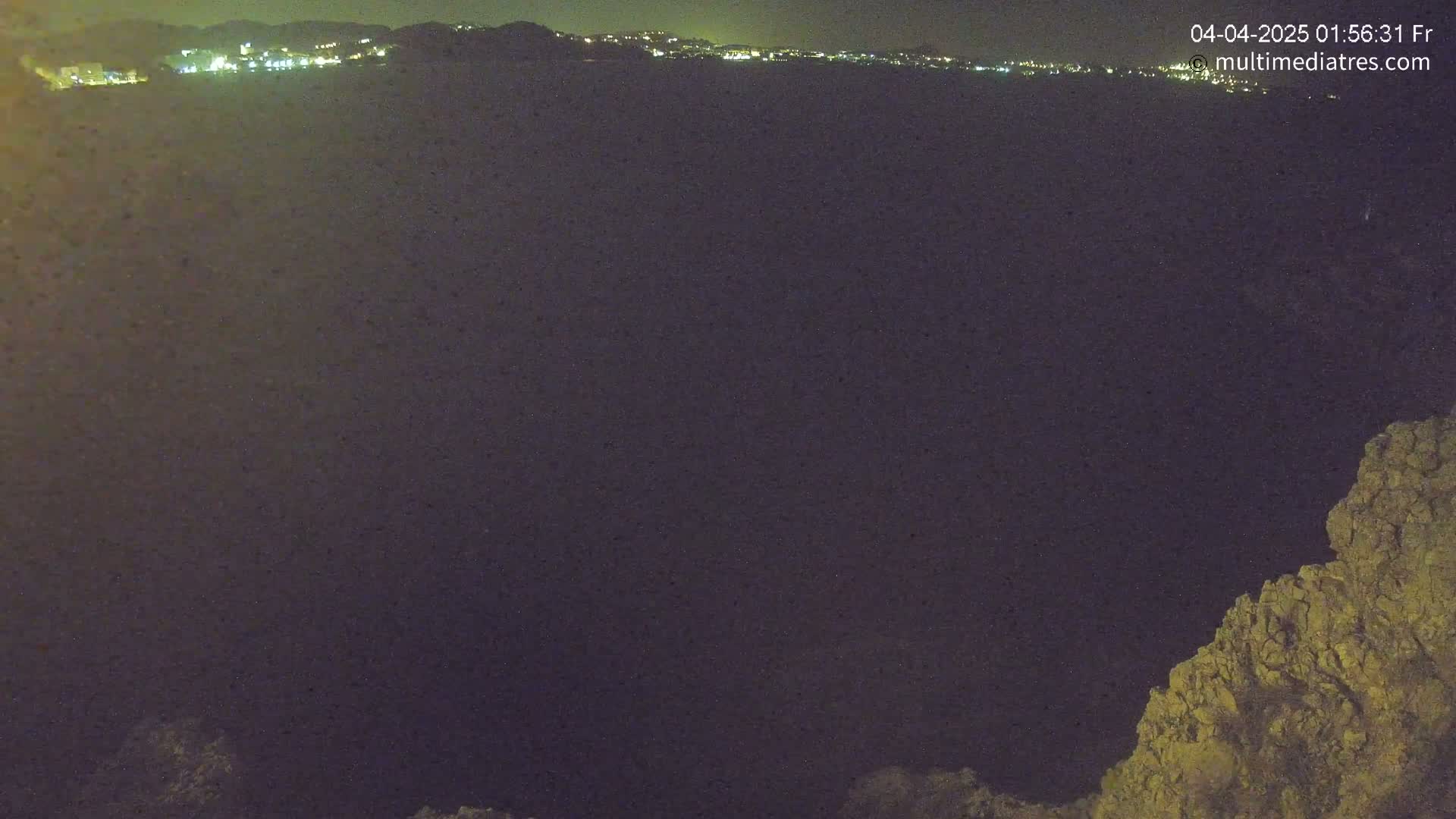 A nighttime view of a dark body of water with a distant, illuminated cityscape and a rocky outcrop in the foreground under a clear, dark sky.
