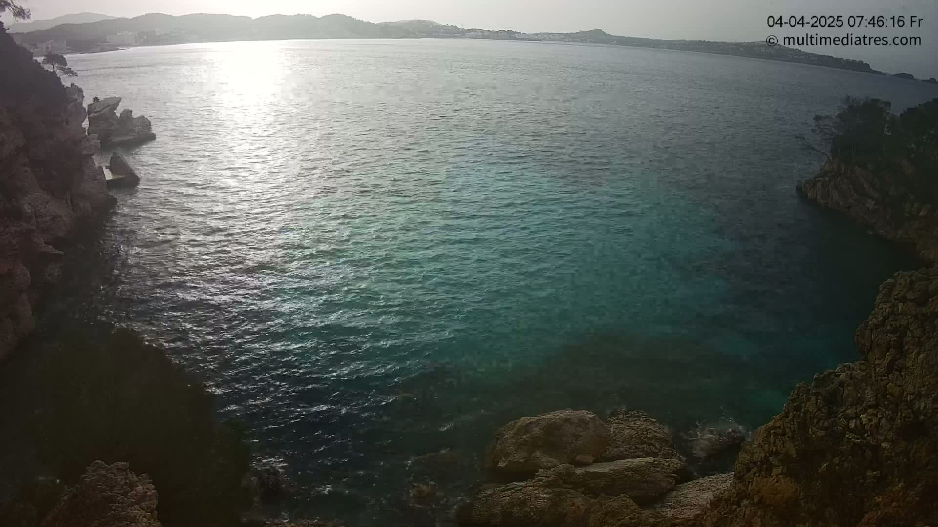 A tranquil cove of dark teal water is nestled between rocky cliffs, under a hazy, sunlit sky.