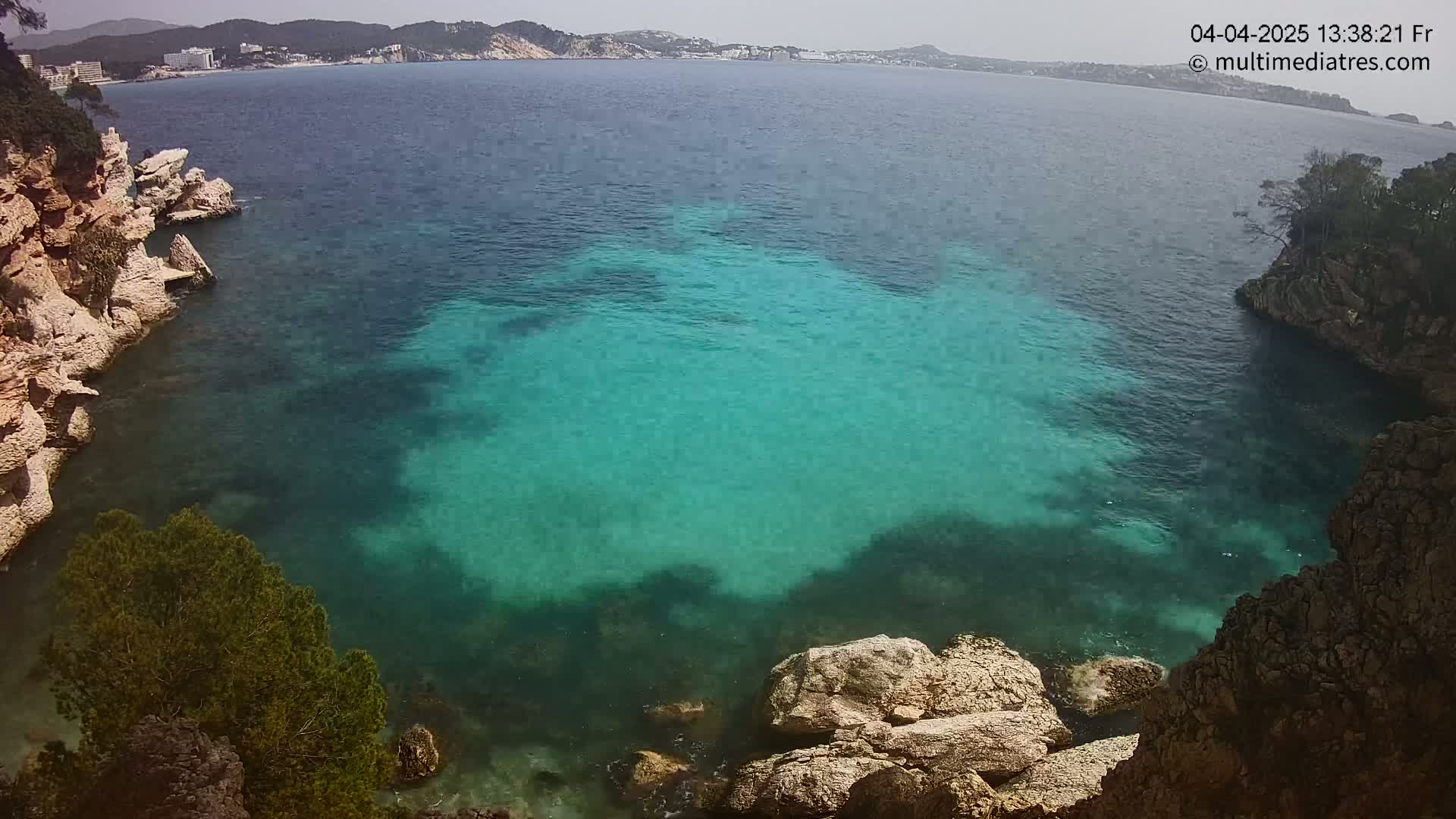 A cove of clear, teal water is nestled between light-colored rocky cliffs on a sunny day, with a distant coastline visible in the background.