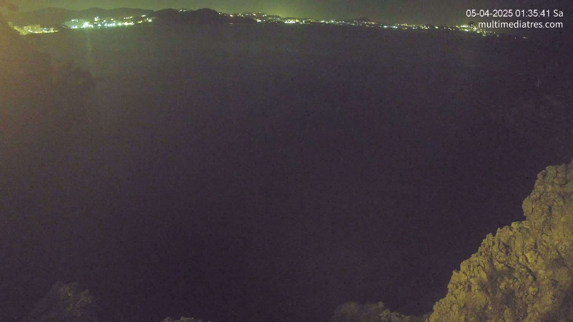 A nighttime, dark view of a body of water with a distant, illuminated town in the background and a rocky outcrop in the foreground.