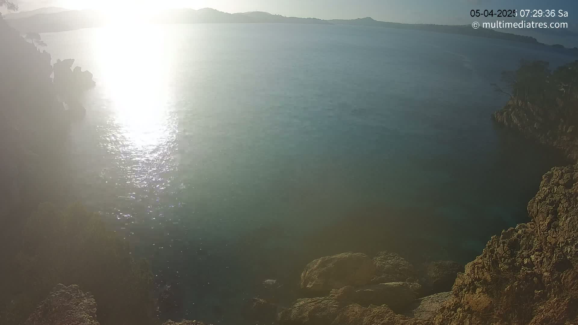 Rocky shores surround a calm body of water reflecting bright sunlight in a hazy, sunny morning.