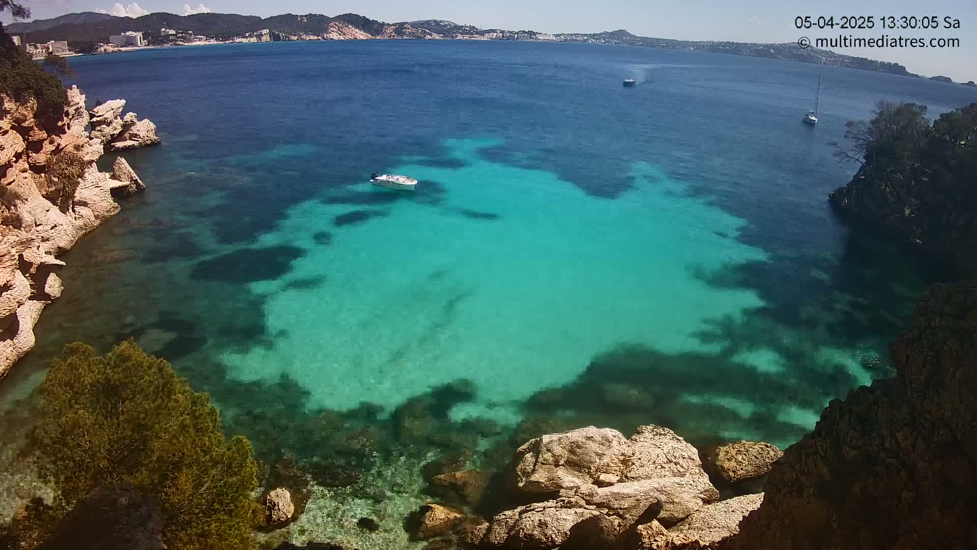 A sunny day shows a clear, turquoise cove surrounded by light-brown rocks and green vegetation, with several small boats visible on the calm, blue water beyond.