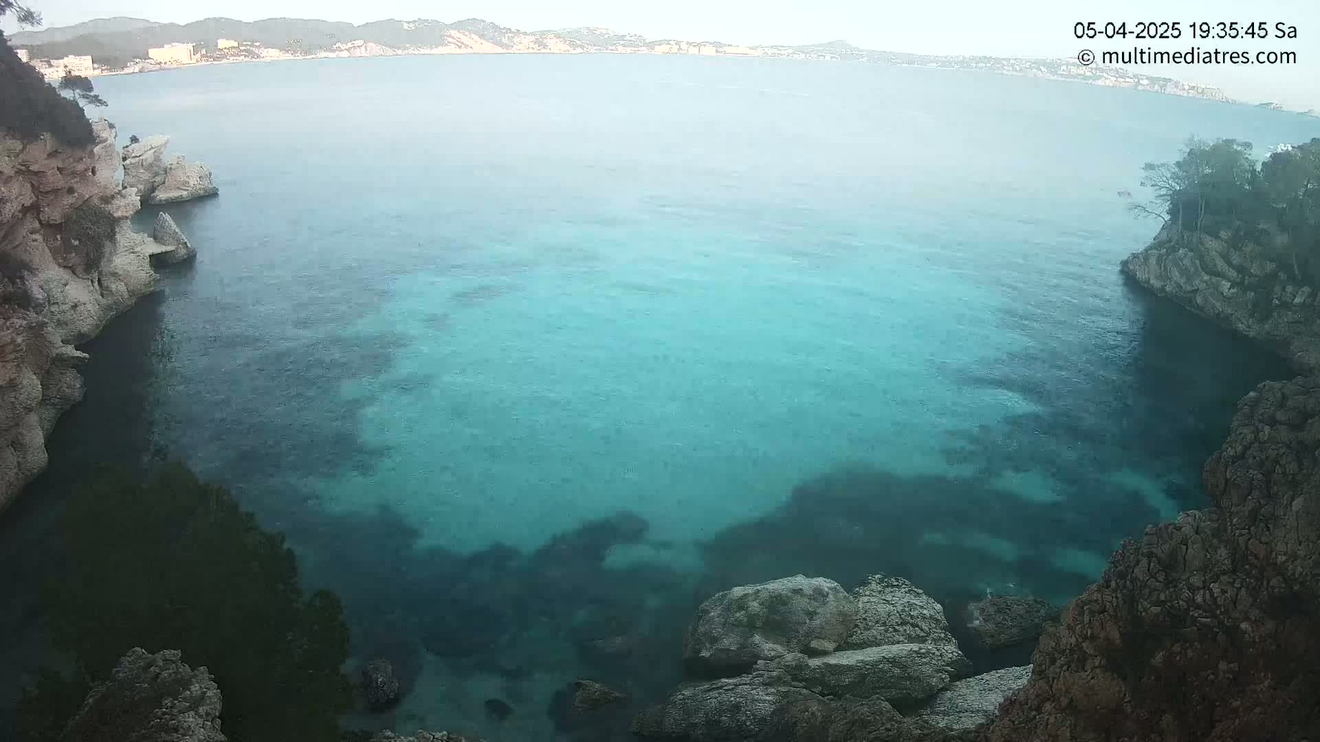 Clear, teal water fills a cove between rocky, vegetated cliffs on a partly sunny day.