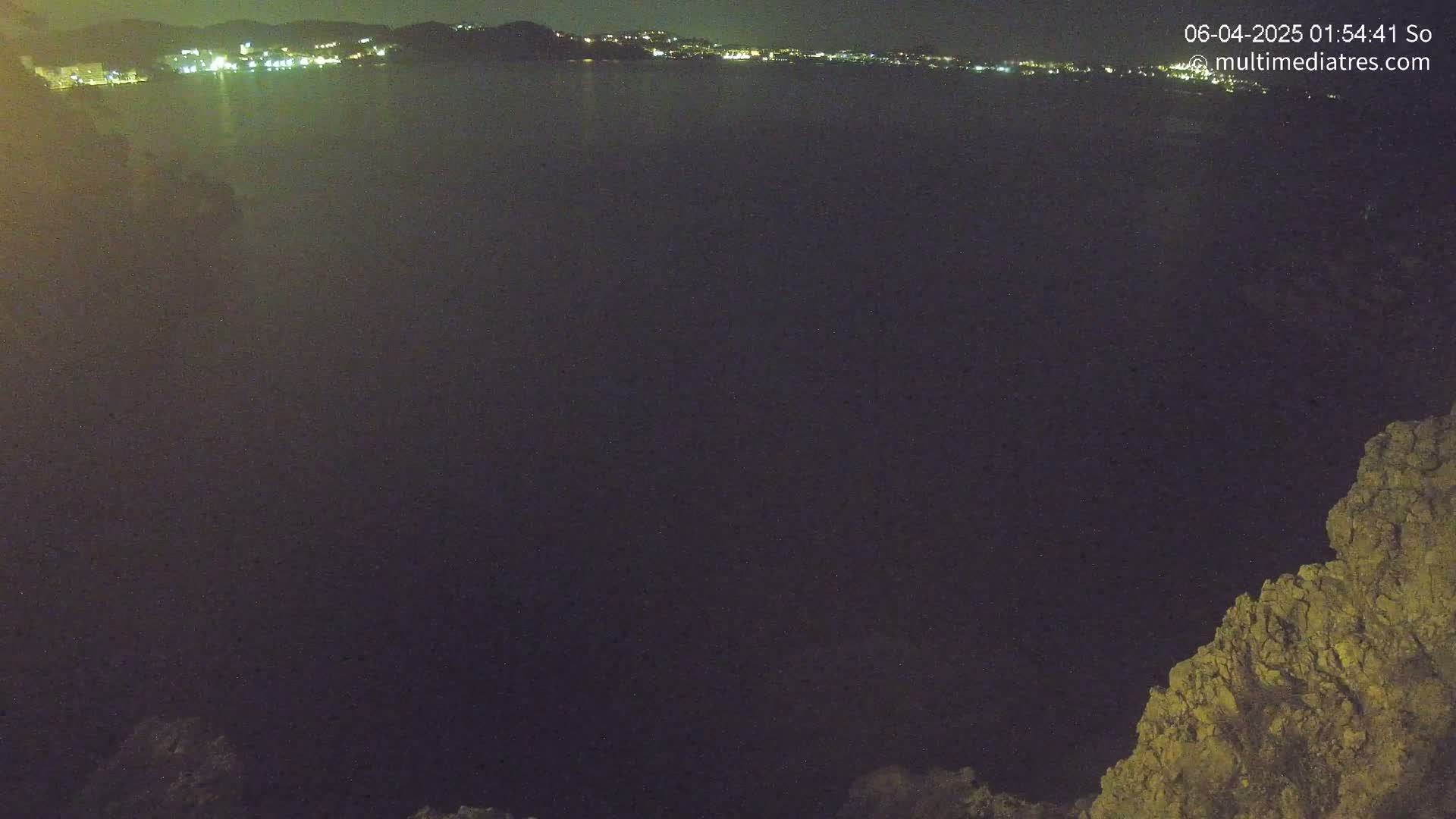 A nighttime view of a dark body of water with a distant, illuminated cityscape and a rocky outcrop in the lower right corner.