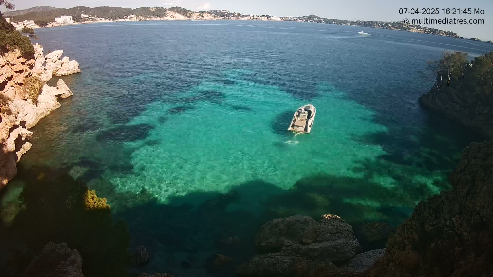 A motorboat floats in clear, teal water near rocky shorelines on a sunny day.