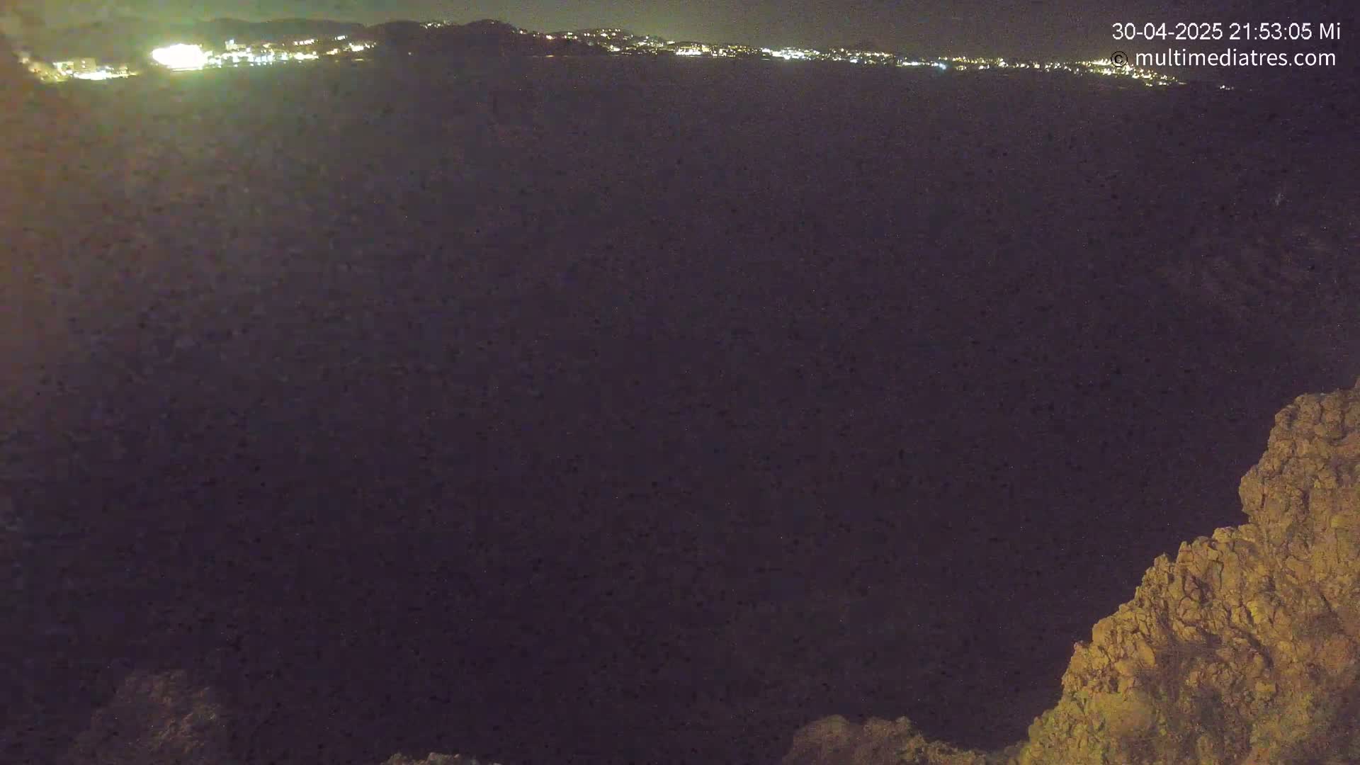 A nighttime view shows a distant, illuminated city across a dark body of water, with a rocky outcrop in the lower right corner.
