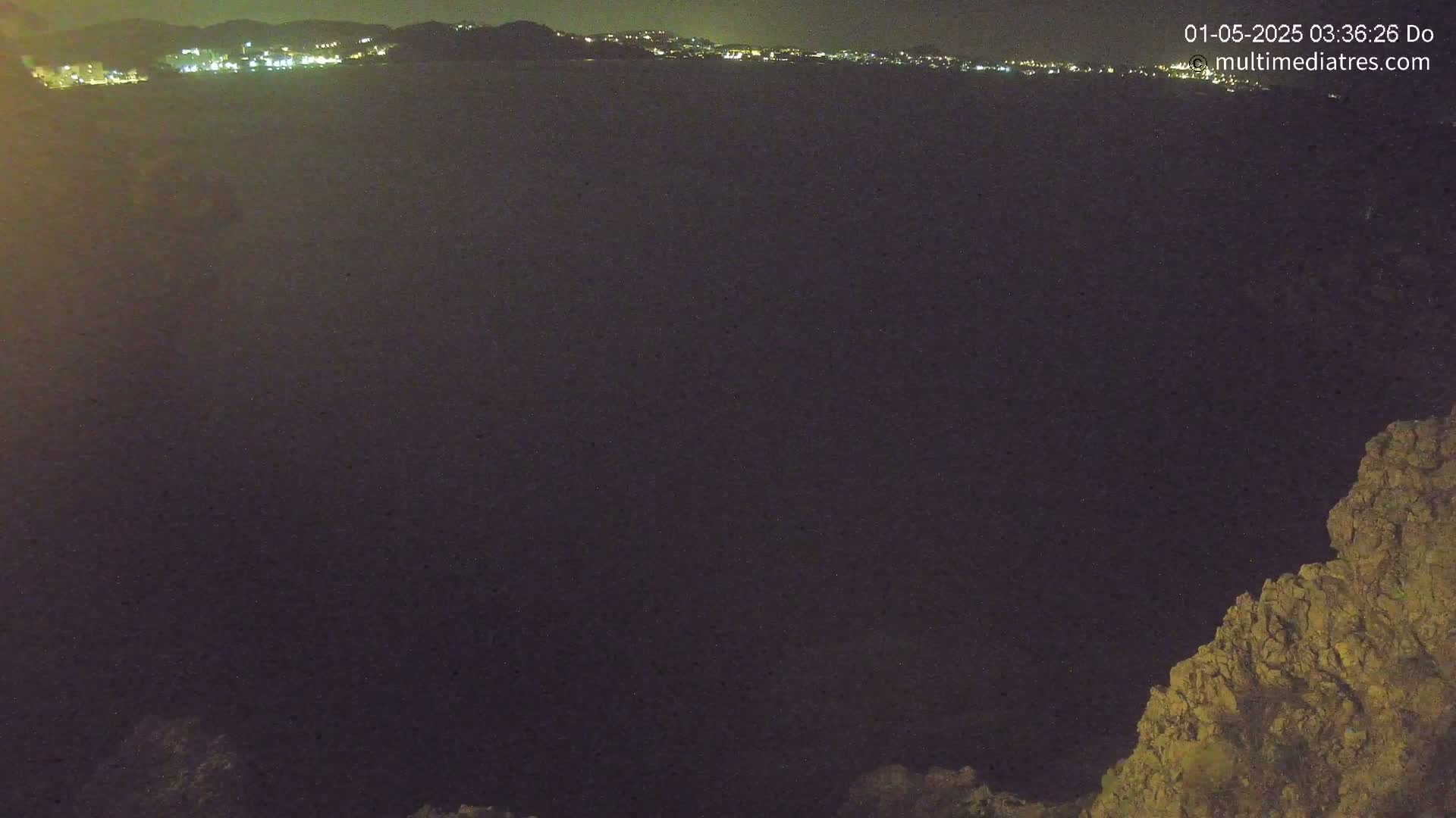 A nighttime view shows a dark body of water with a distant city skyline illuminated, and a rocky outcrop in the lower right corner.