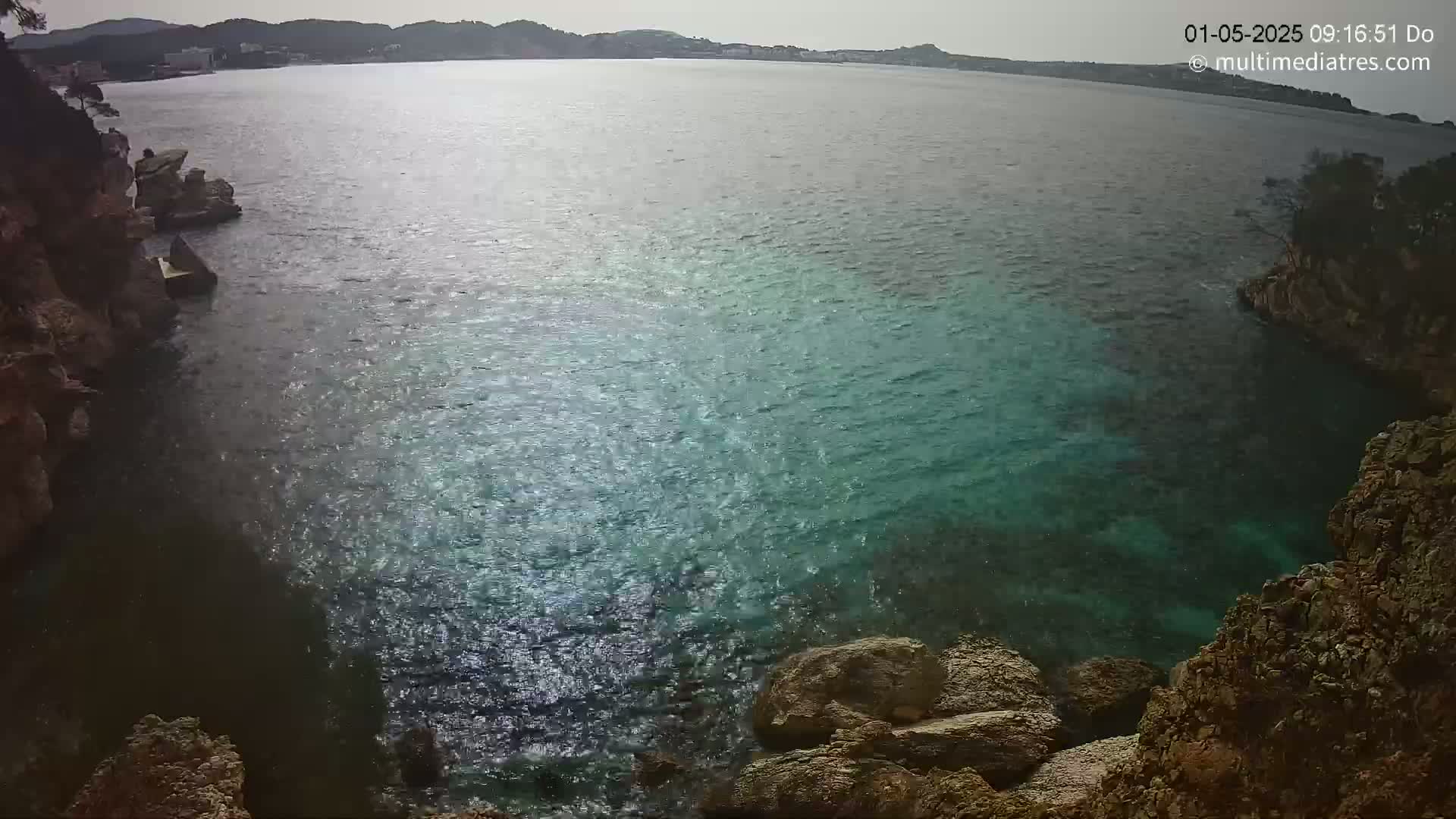 A calm, clear body of water is seen from a high vantage point overlooking rocky, cliff-like formations on either side, with a distant shoreline visible in the background under a somewhat overcast sky.