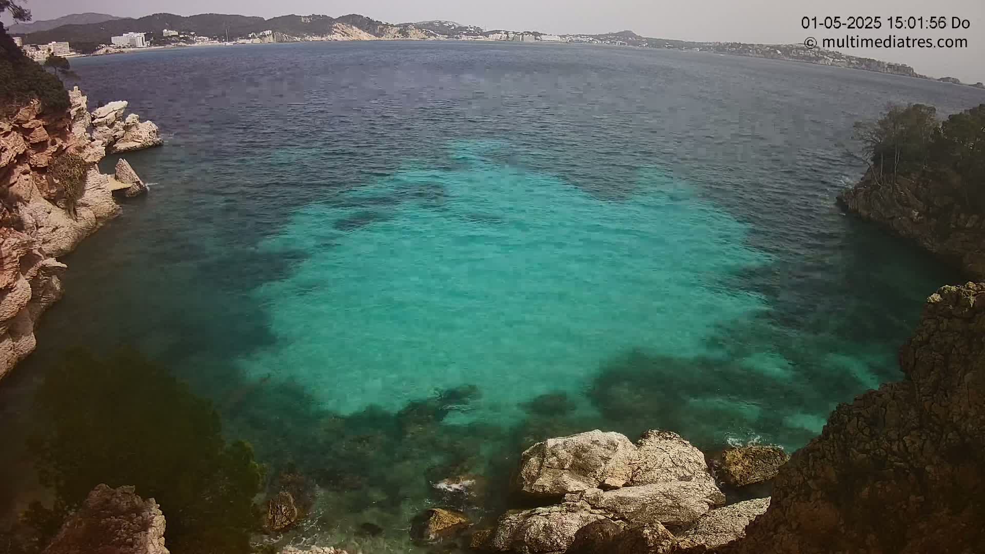 A high-angle view shows a cove of clear, teal water nestled between rocky, tan cliffs on a partly cloudy day.