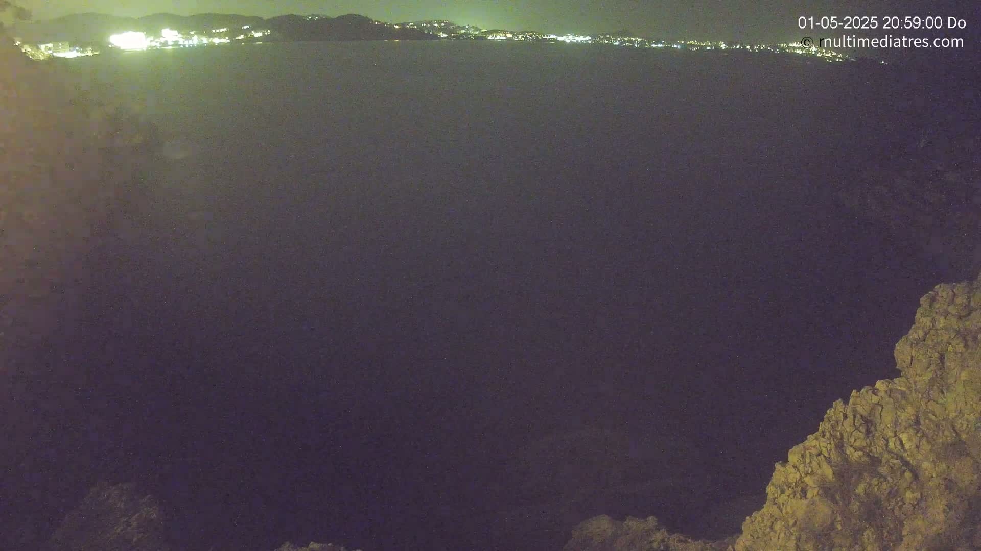 A nighttime view of a dark body of water with a distant, faintly lit cityscape and a rocky outcrop in the foreground.