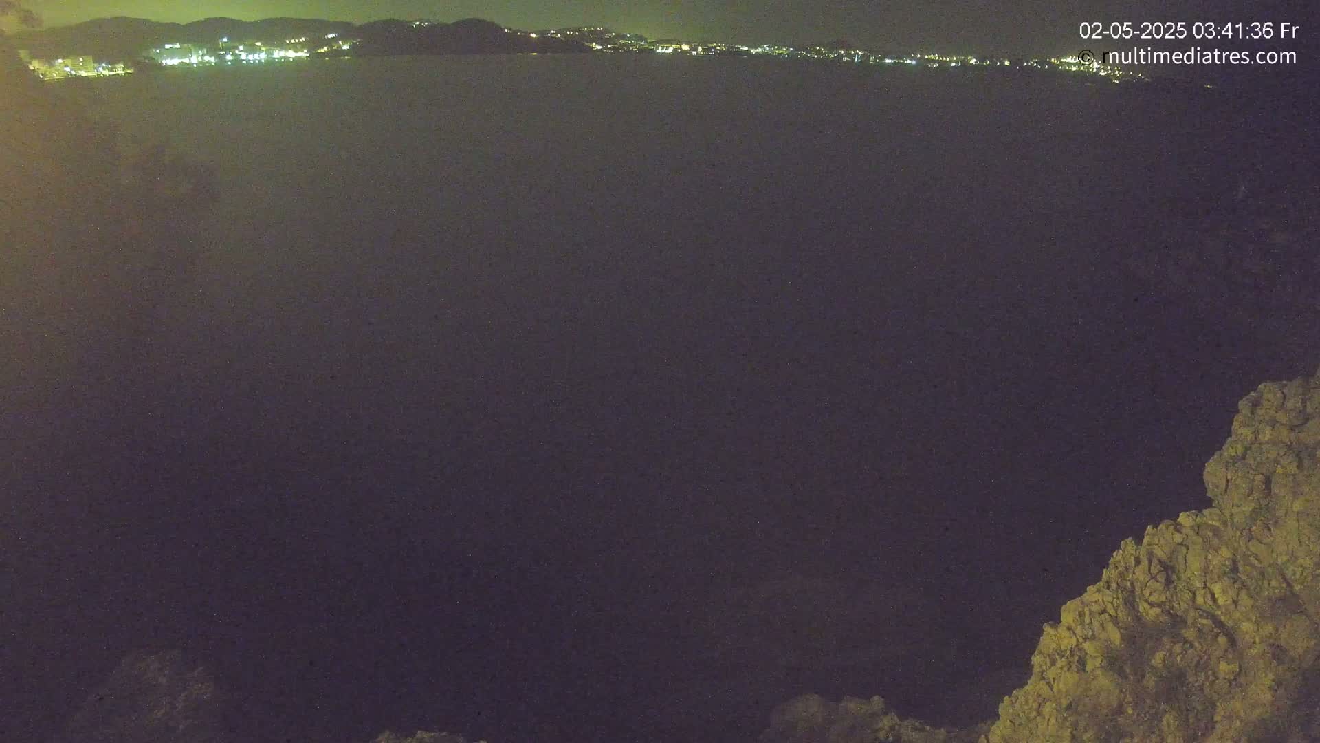 A nighttime view of a dark body of water with a distant, illuminated cityscape and a rocky outcrop in the lower right corner.