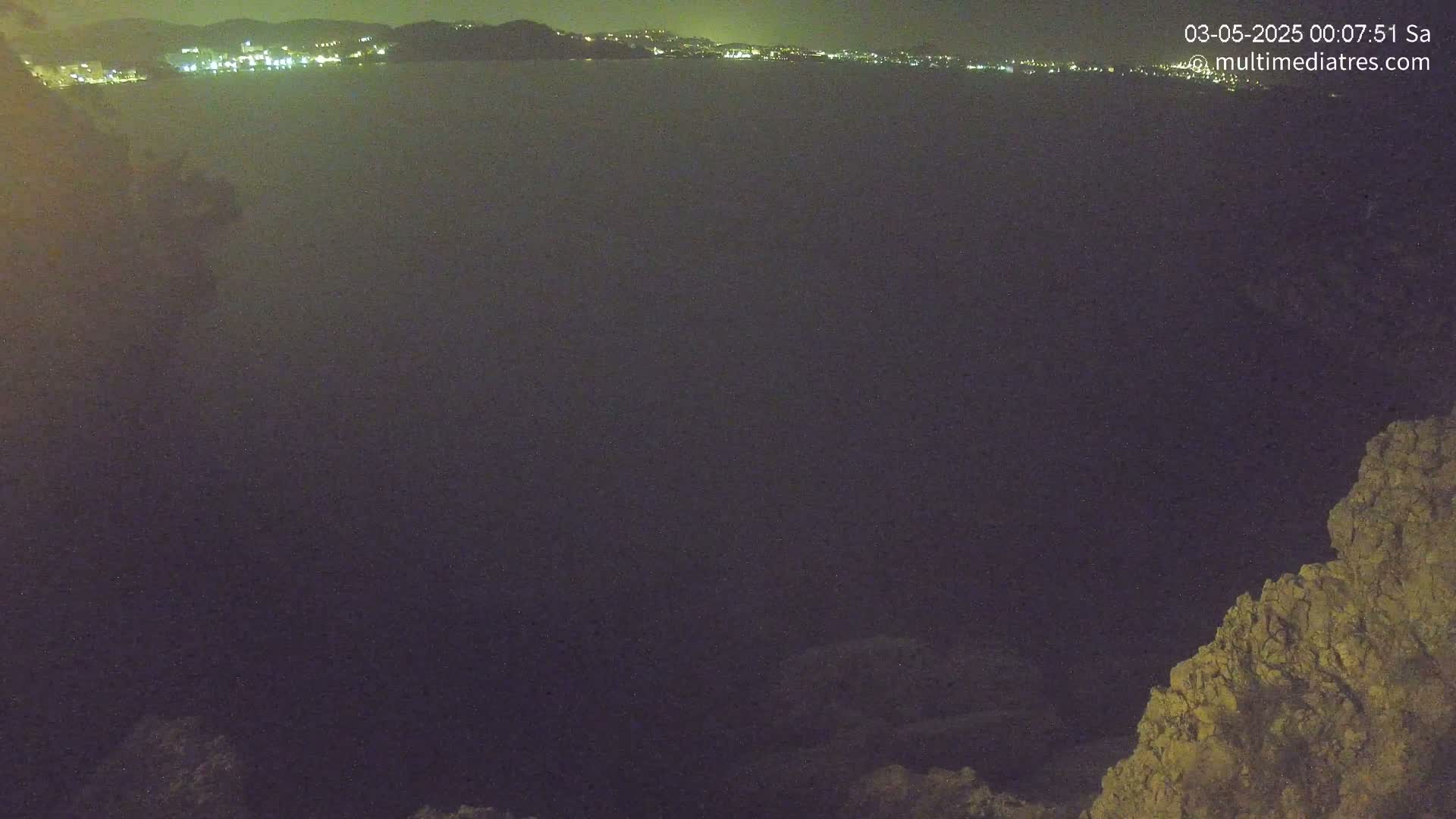 A dark, nighttime view shows a calm body of water with a distant, faintly lit coastline and rocky terrain in the foreground.