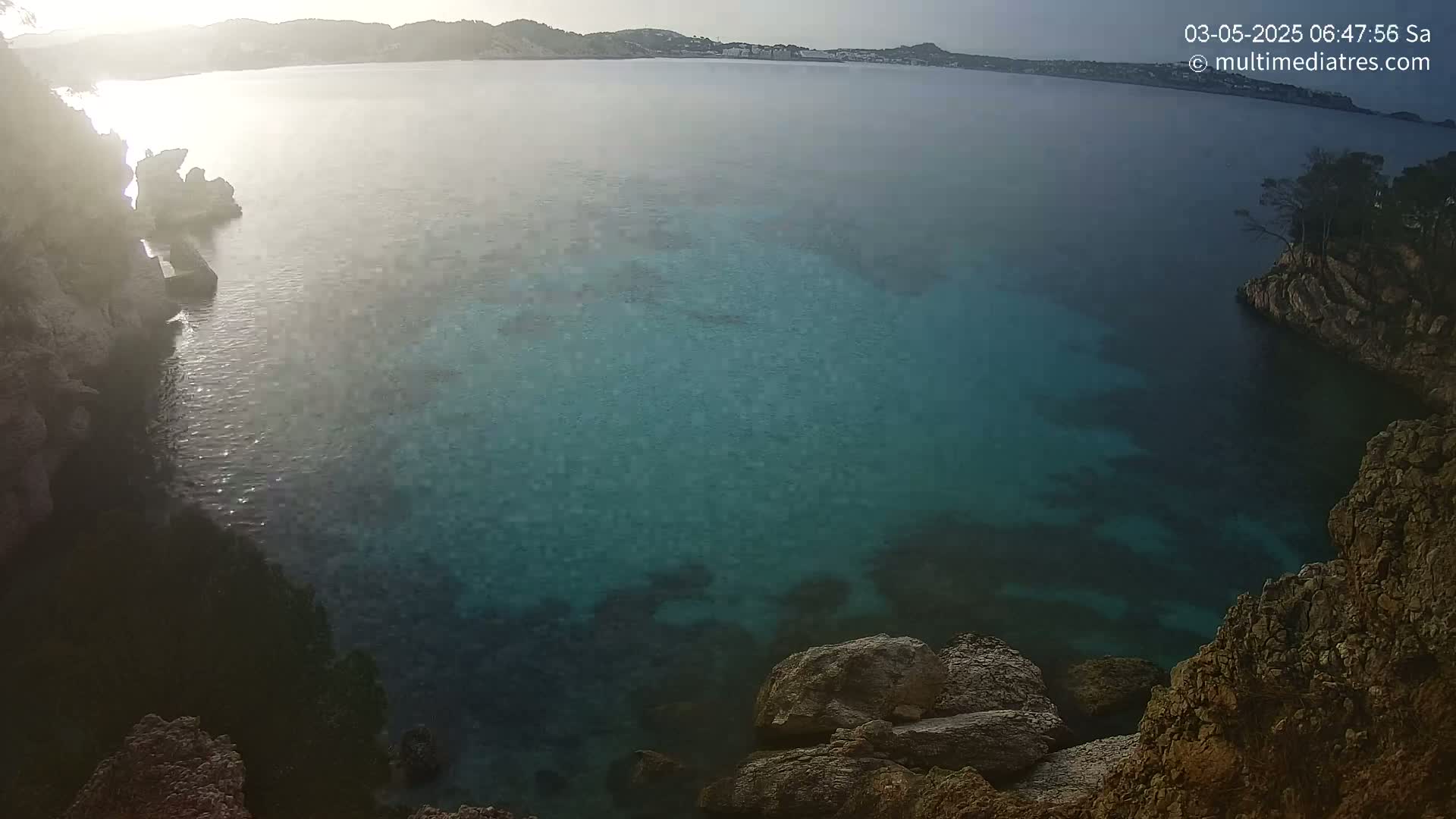 A tranquil cove of clear, teal water is surrounded by rocky cliffs under a bright, possibly sunrise, sky.