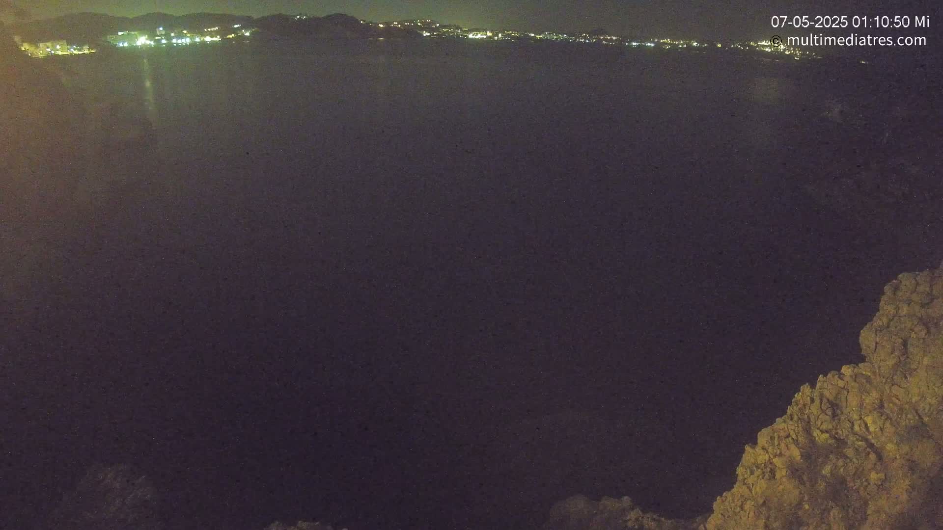 A nighttime view of a dark body of water with a distant, faintly lit coastline and a rocky outcrop in the foreground.