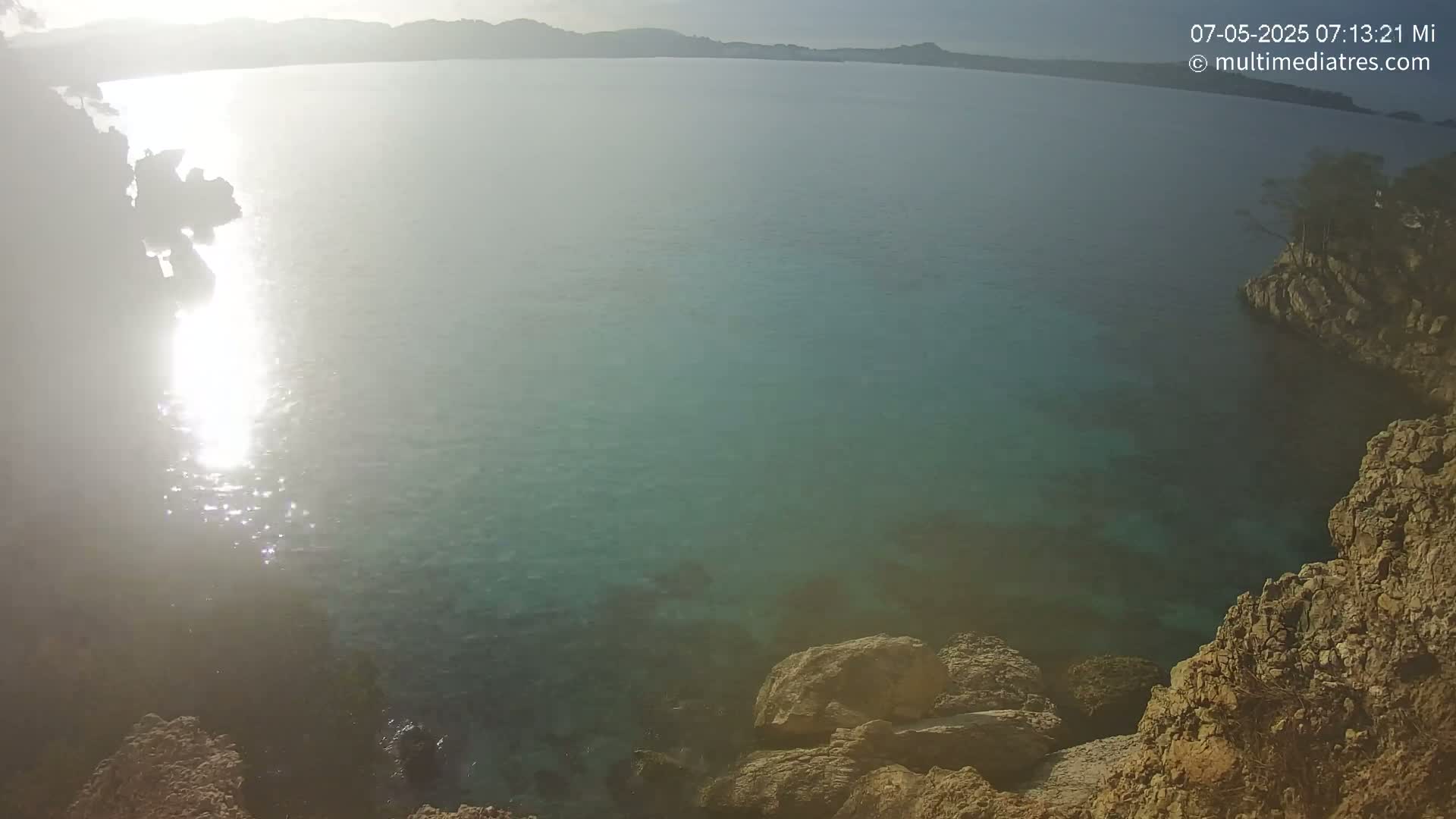 Rocky shores meet a calm, clear turquoise sea under a bright, sunny sky.