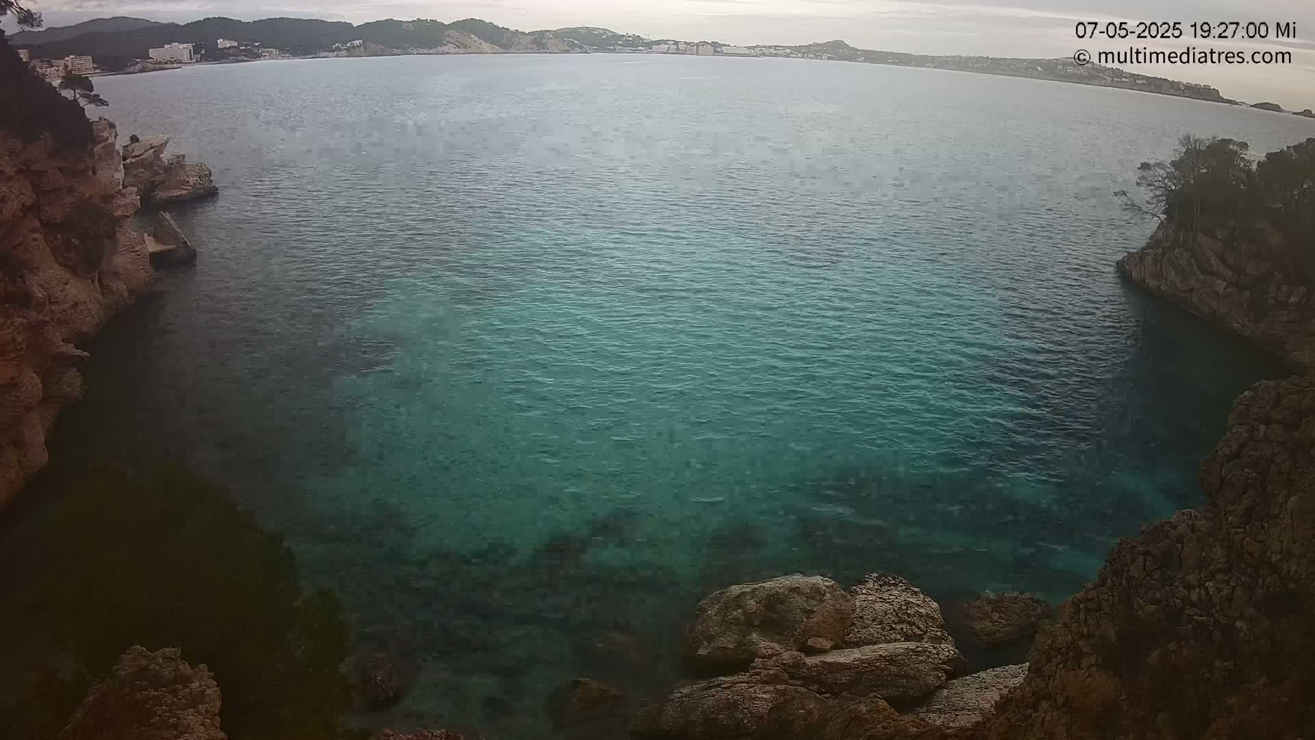 A tranquil cove of teal water is nestled between rocky, cliff-like shores under an overcast sky.