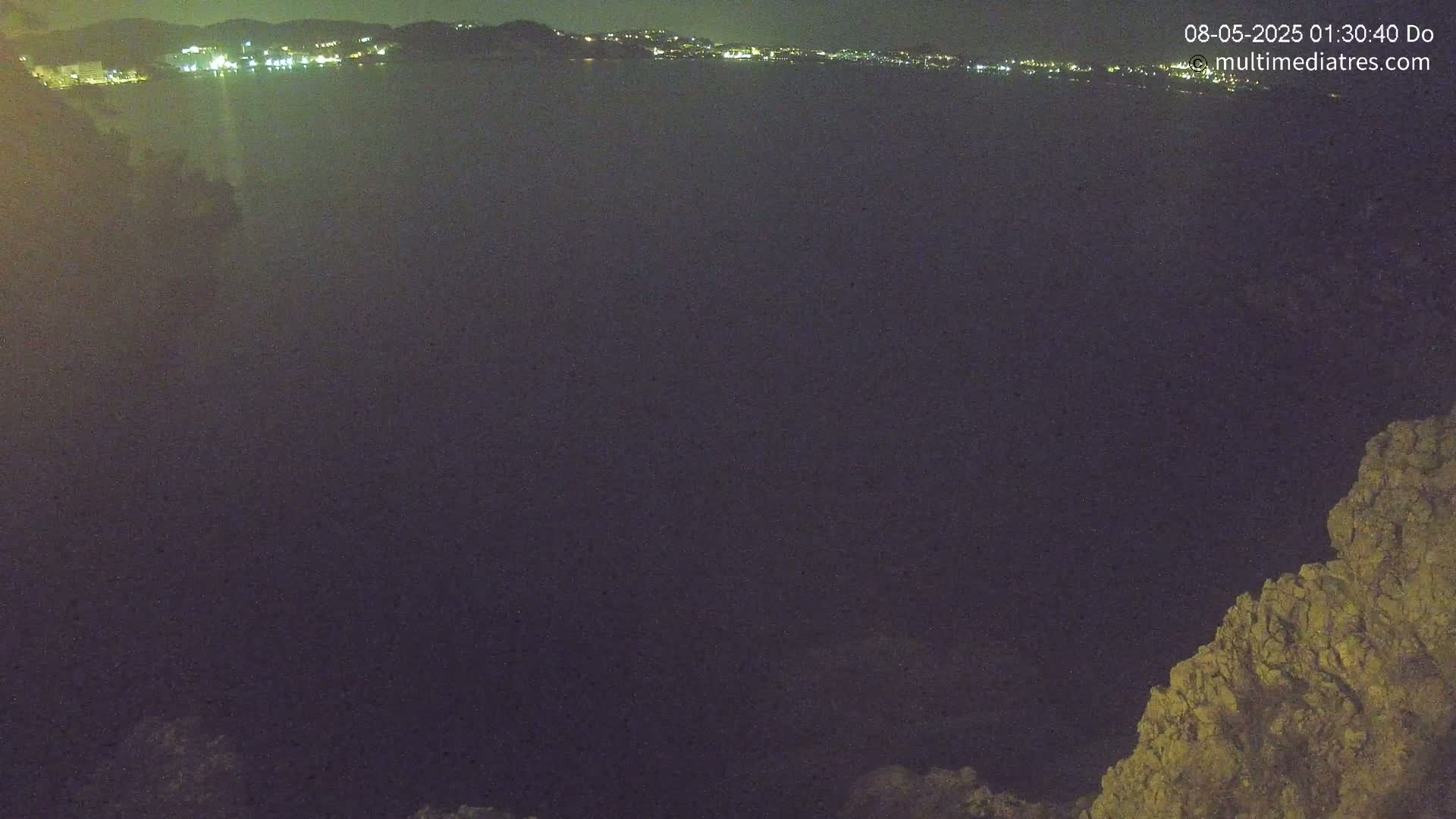 A nighttime view shows a dark body of water with a distant line of city lights, and a rocky outcrop in the foreground under a clear, dark sky.