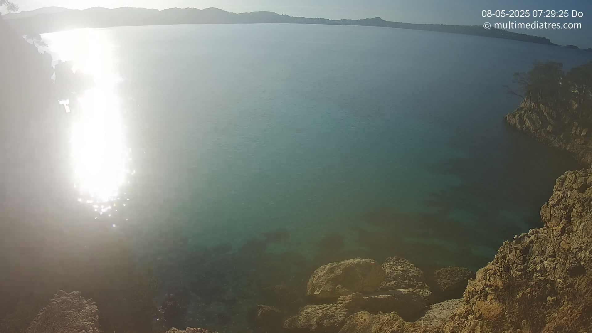 Sunlight glints on the surface of a calm, teal-colored sea, partially enclosed by rocky shores under a clear sky.