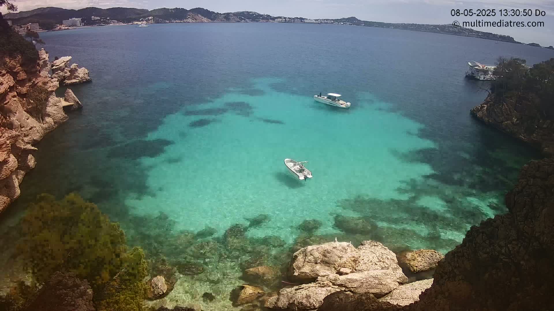 A tranquil cove of clear turquoise water, nestled between rocky cliffs, with two small boats at anchor under a sunny sky.
