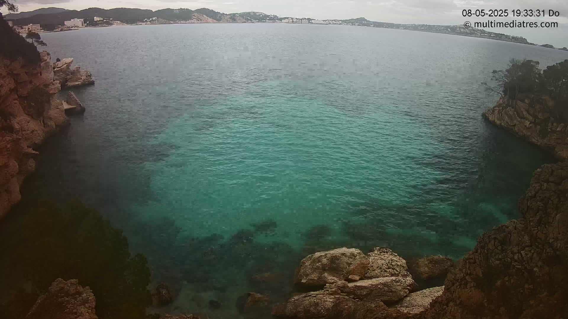 A cove of teal water is nestled between rocky cliffs on an overcast day, with a distant shoreline visible in the background.