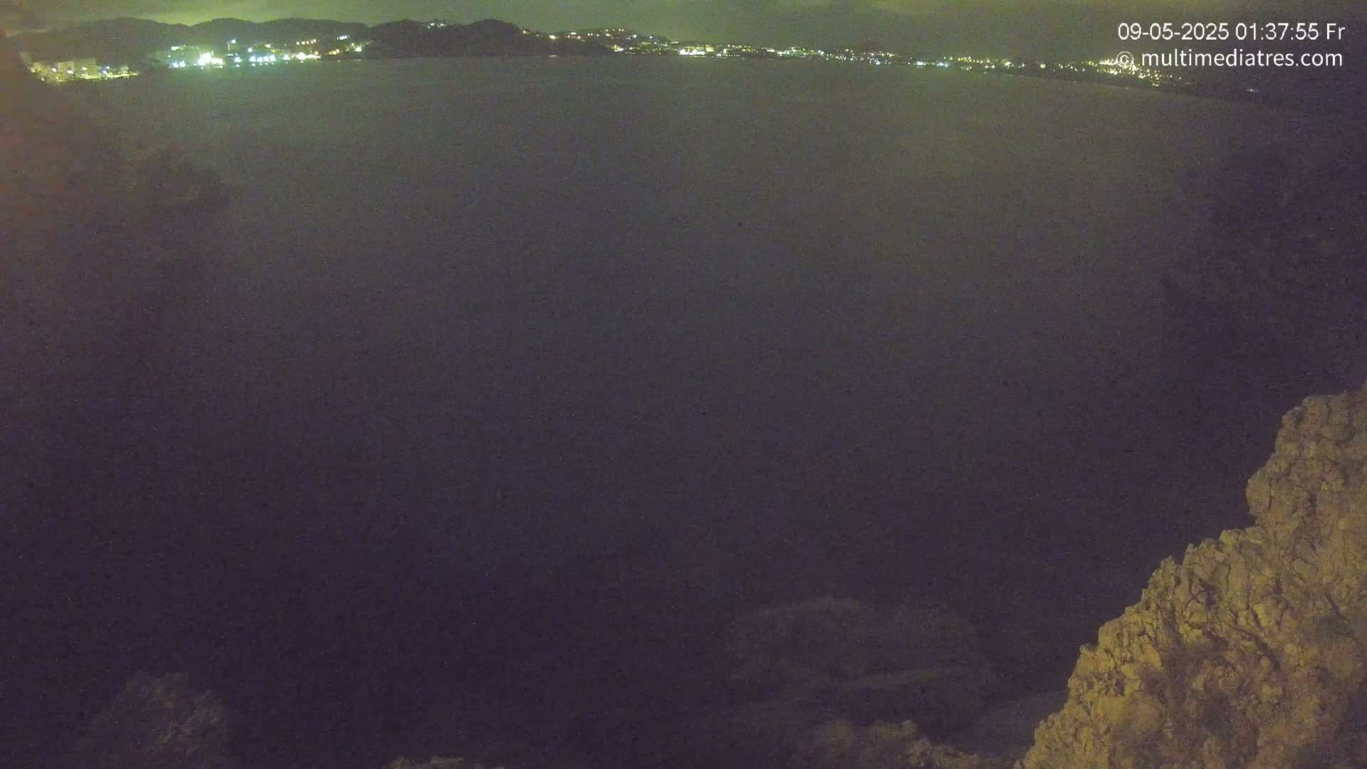 A nighttime view shows a dark body of water with a distant, faintly lit cityscape under a clear, dark sky, partially obscured by a rocky outcrop in the lower right corner.