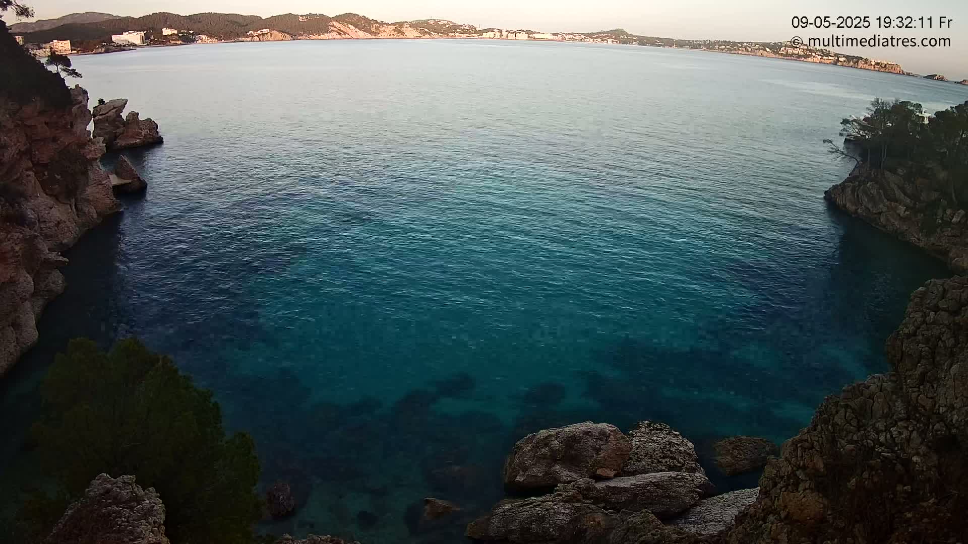 Dark teal water fills a cove between rocky, tree-covered shores, with a distant coastline visible under a calm sky.