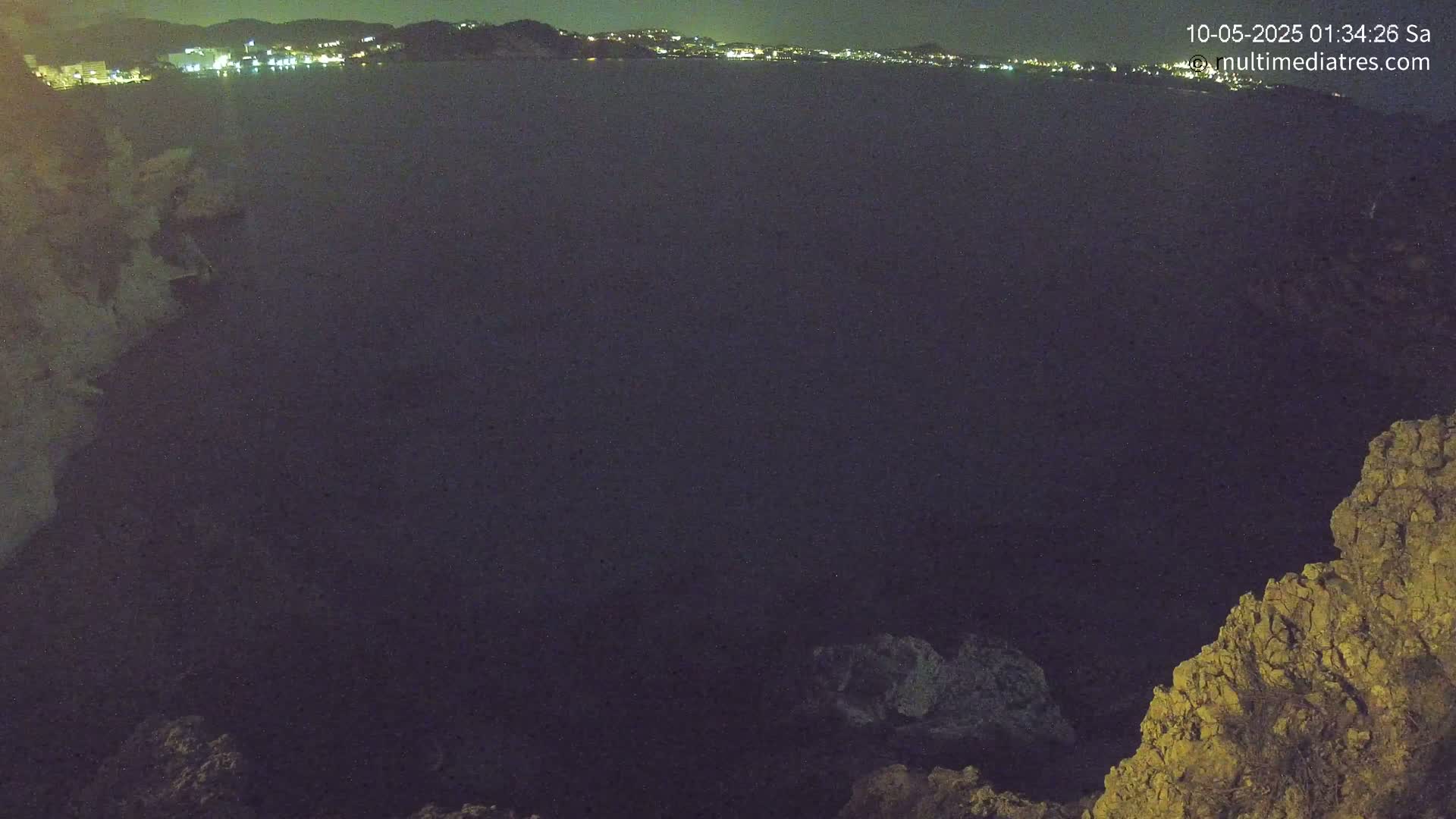 A nighttime view of a dark body of water with a distant city skyline illuminated, seen from a rocky outcrop.