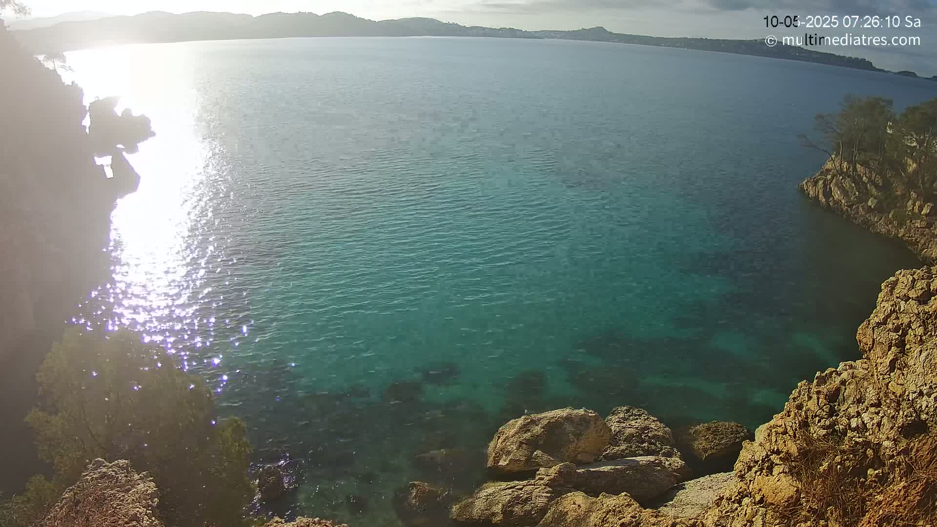 Sunlight glistens on the surface of a calm, teal-colored sea, partially enclosed by rocky, tan-colored cliffs under a partly cloudy sky.