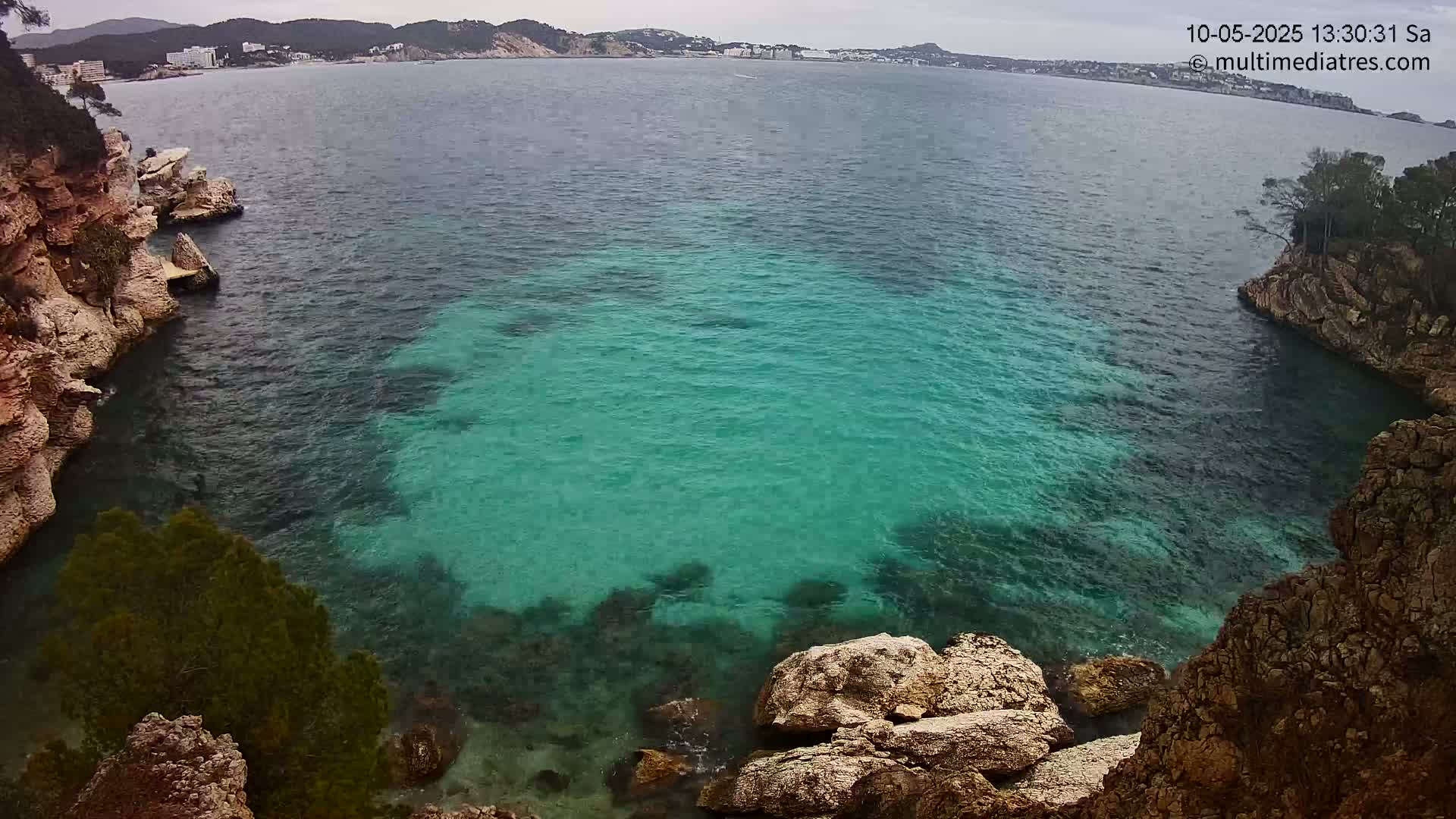 A cove of clear, teal water is nestled between rocky cliffs on an overcast day, with a distant town visible across the water.