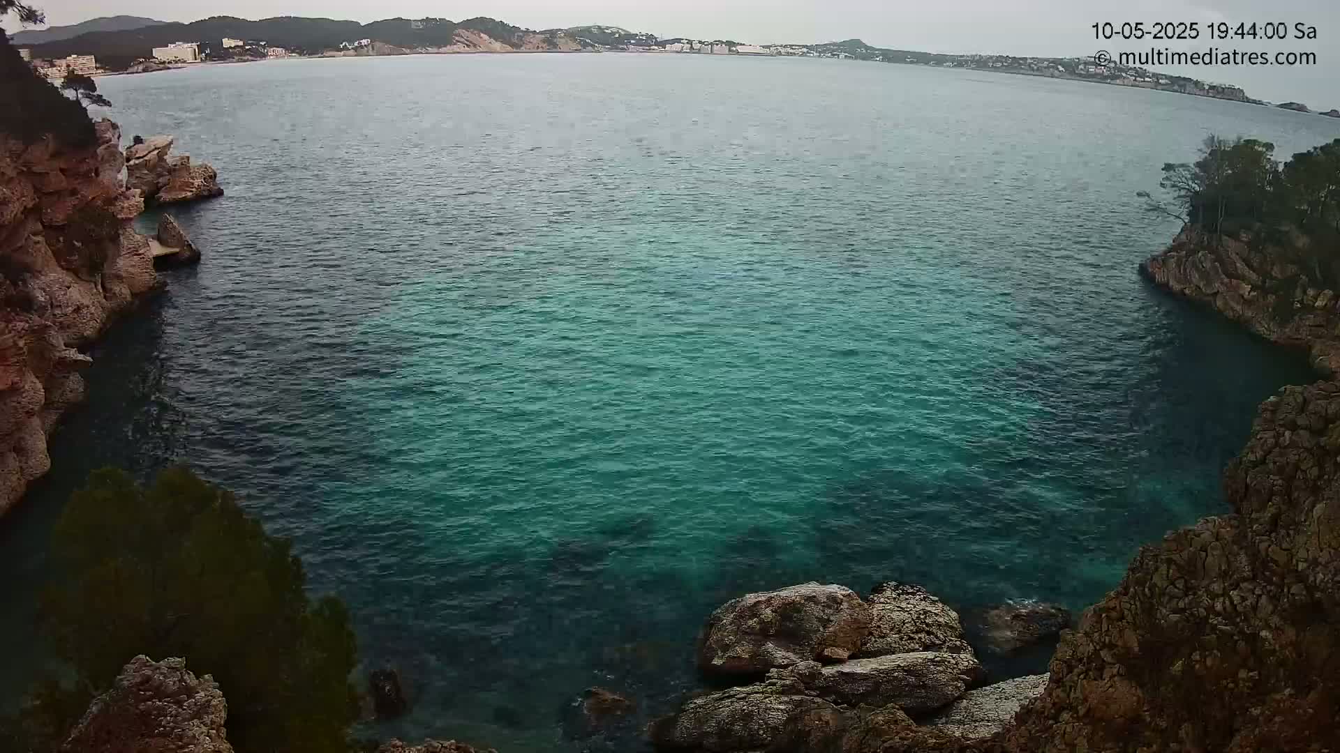 A tranquil teal cove is nestled between rocky cliffs, with a distant coastline visible under an overcast sky.