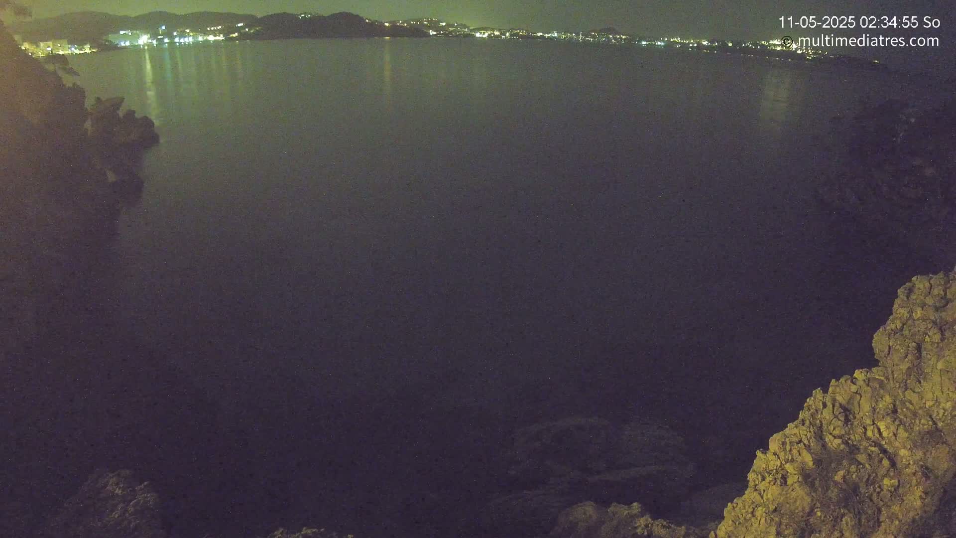 A nighttime, wide shot of a dark body of water with a distant, faintly lit town in the background and rocky terrain in the foreground.