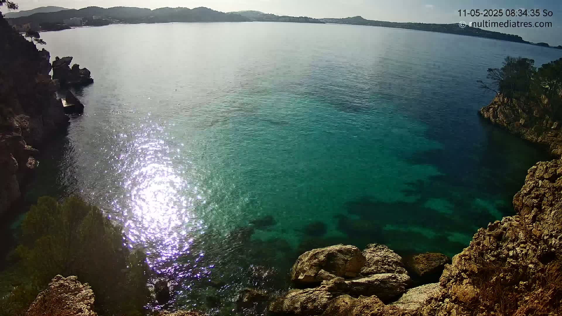 Sunlight glistens on a calm, teal sea nestled between rocky, vegetated shores.