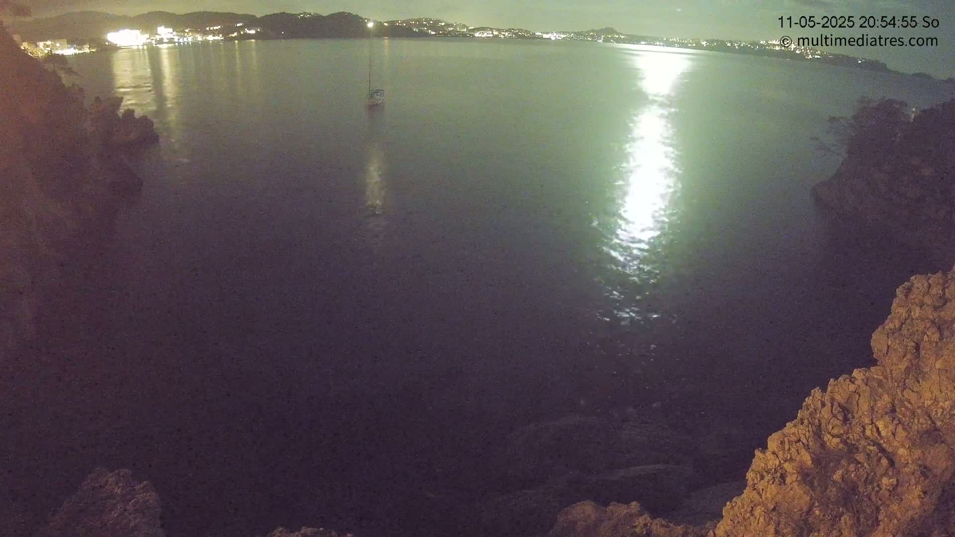A nighttime view from a rocky outcrop shows a calm body of water reflecting bright moonlight, with a small sailboat visible in the distance and lights from a town on the far shore.