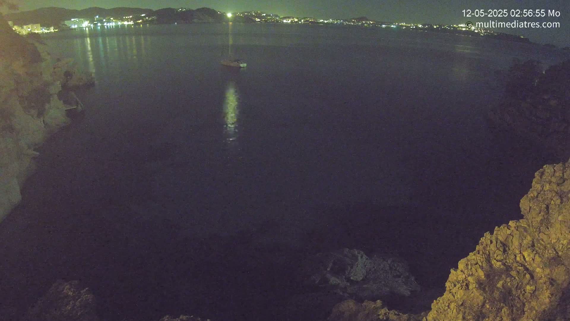 A nighttime view of a dark body of water with a single boat visible, a distant illuminated shoreline, and rocky outcroppings in the foreground.