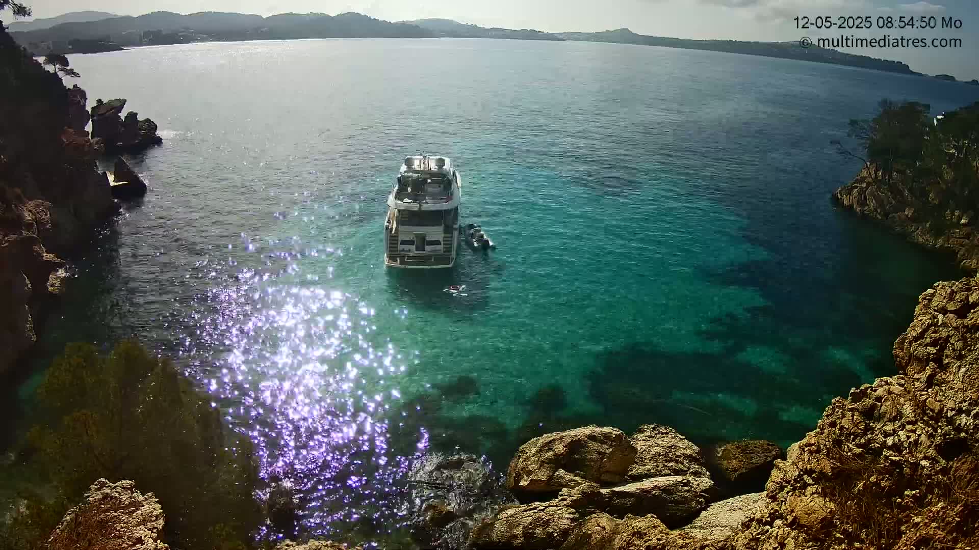 A yacht is anchored in a calm, clear, teal cove surrounded by rocky cliffs on a sunny day.