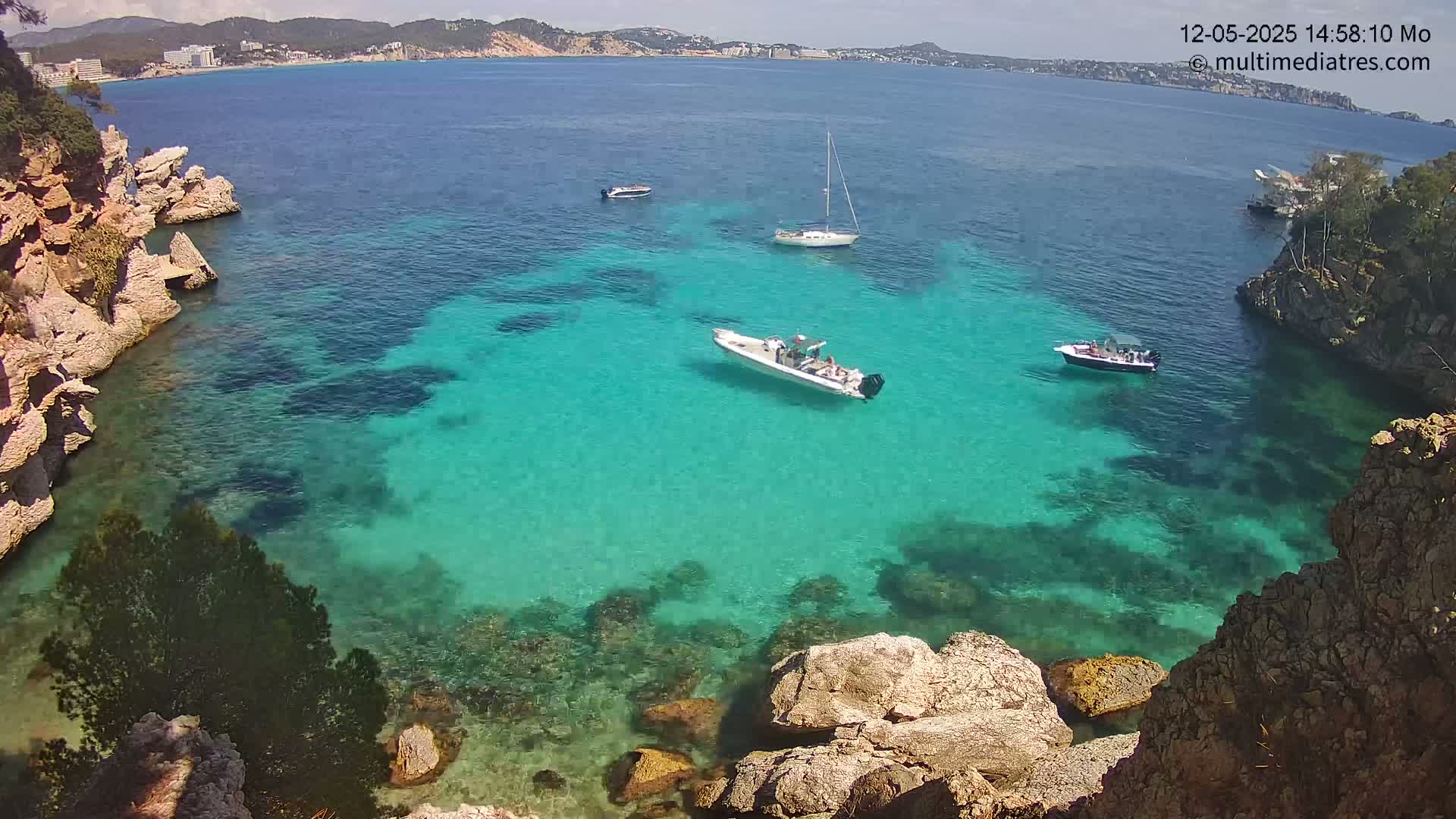 A tranquil cove with crystal-clear turquoise water, surrounded by light-colored rocky cliffs, is dotted with several small boats on a sunny day.