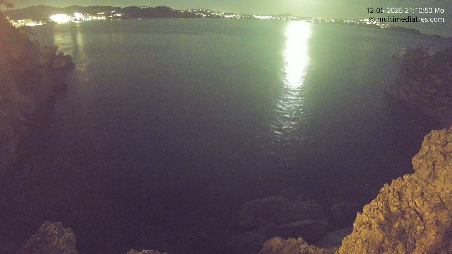 A nighttime view of a calm body of water reflecting city lights on the far shore, seen from a rocky vantage point.