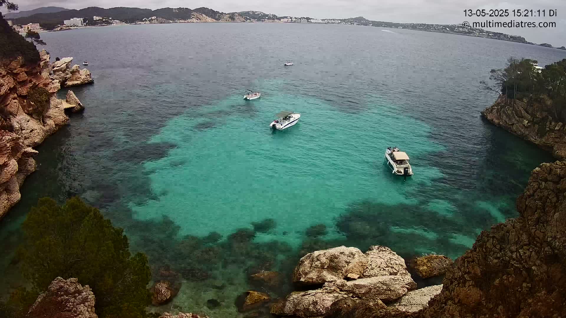 Several boats are anchored in a calm, clear teal cove surrounded by rocky, vegetated cliffs under an overcast sky.
