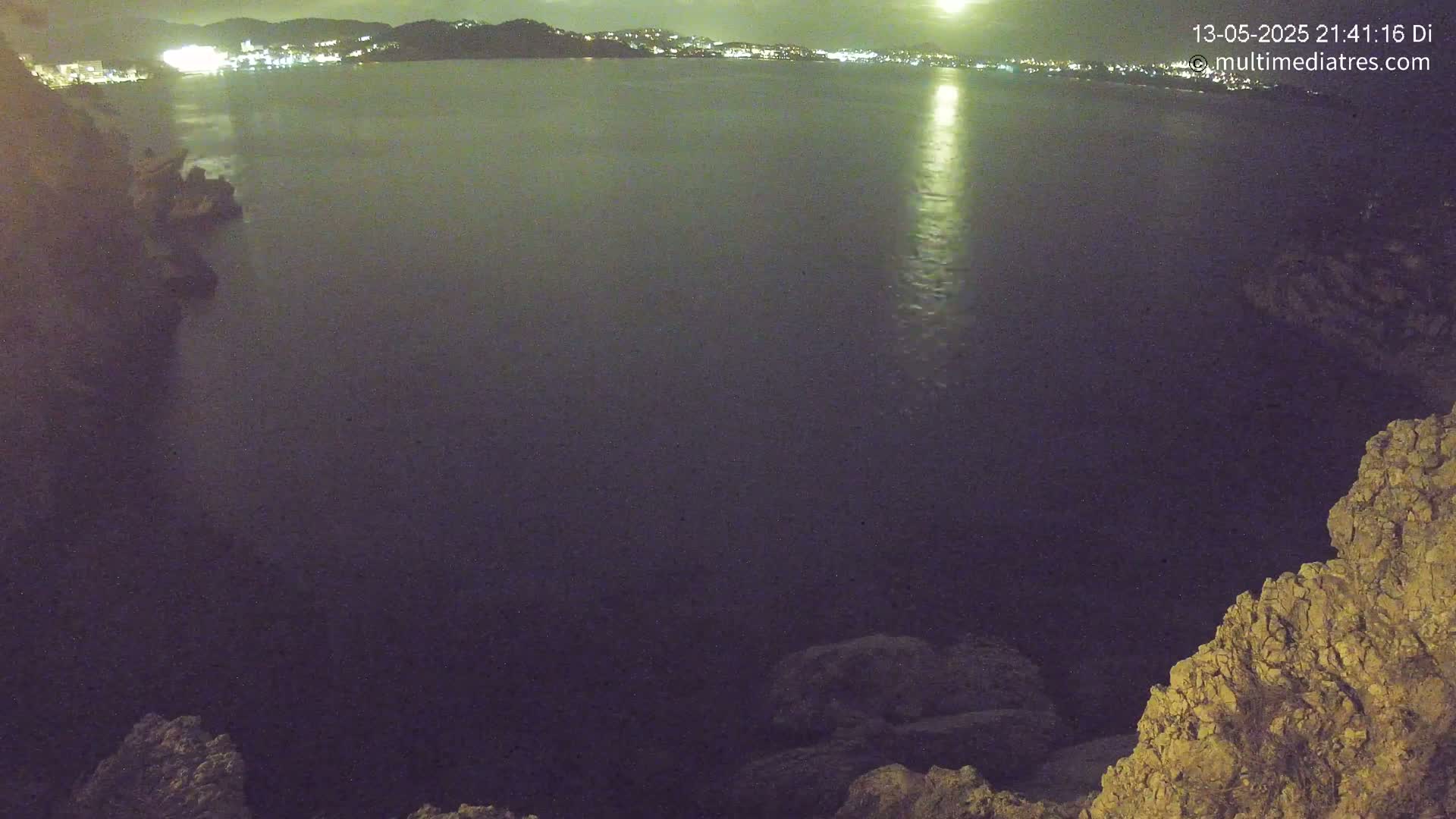A nighttime view of a dark body of water, reflecting city lights on the horizon, from a rocky vantage point.