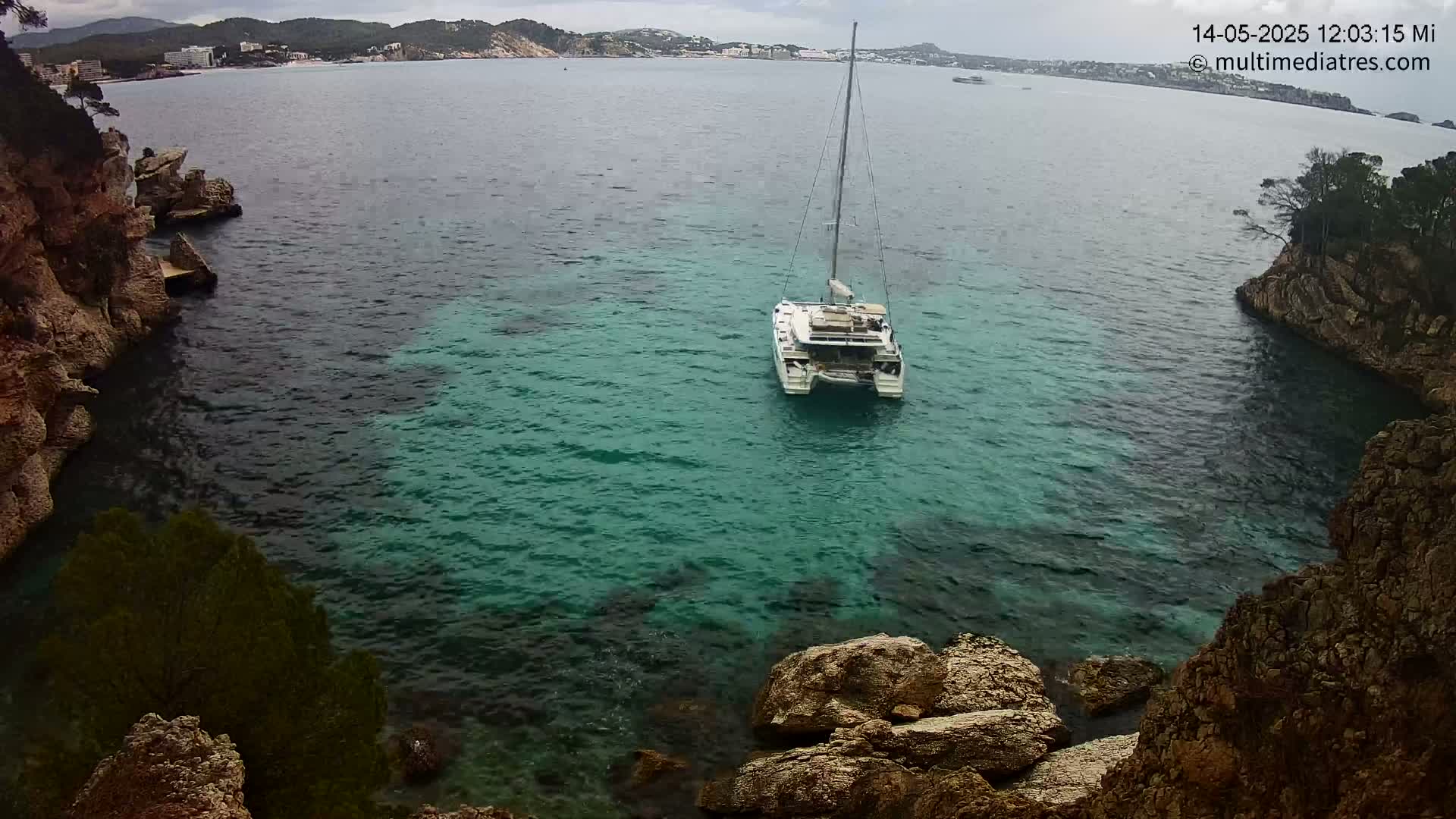 A white catamaran is anchored in a teal cove surrounded by rocky, vegetated cliffs under an overcast sky.