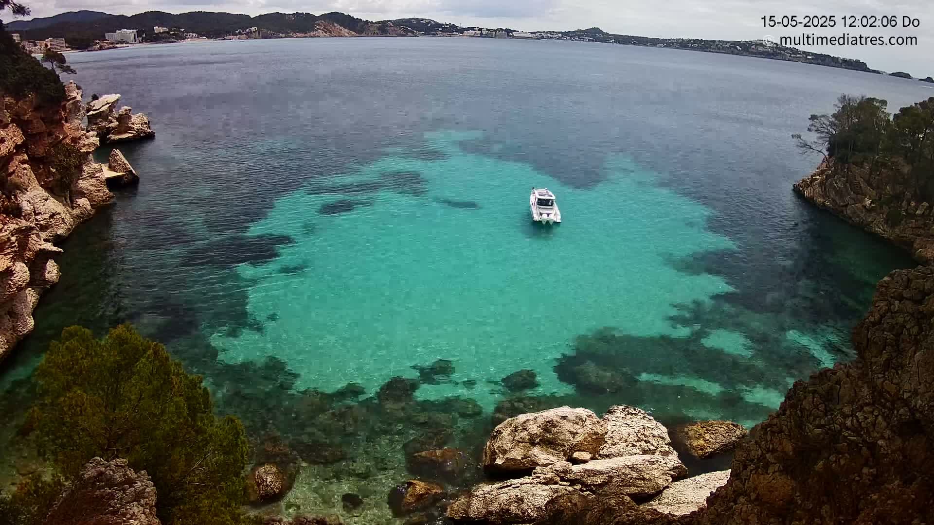 A white boat sits in a calm, clear turquoise cove surrounded by rocky cliffs and vegetation under an overcast sky.