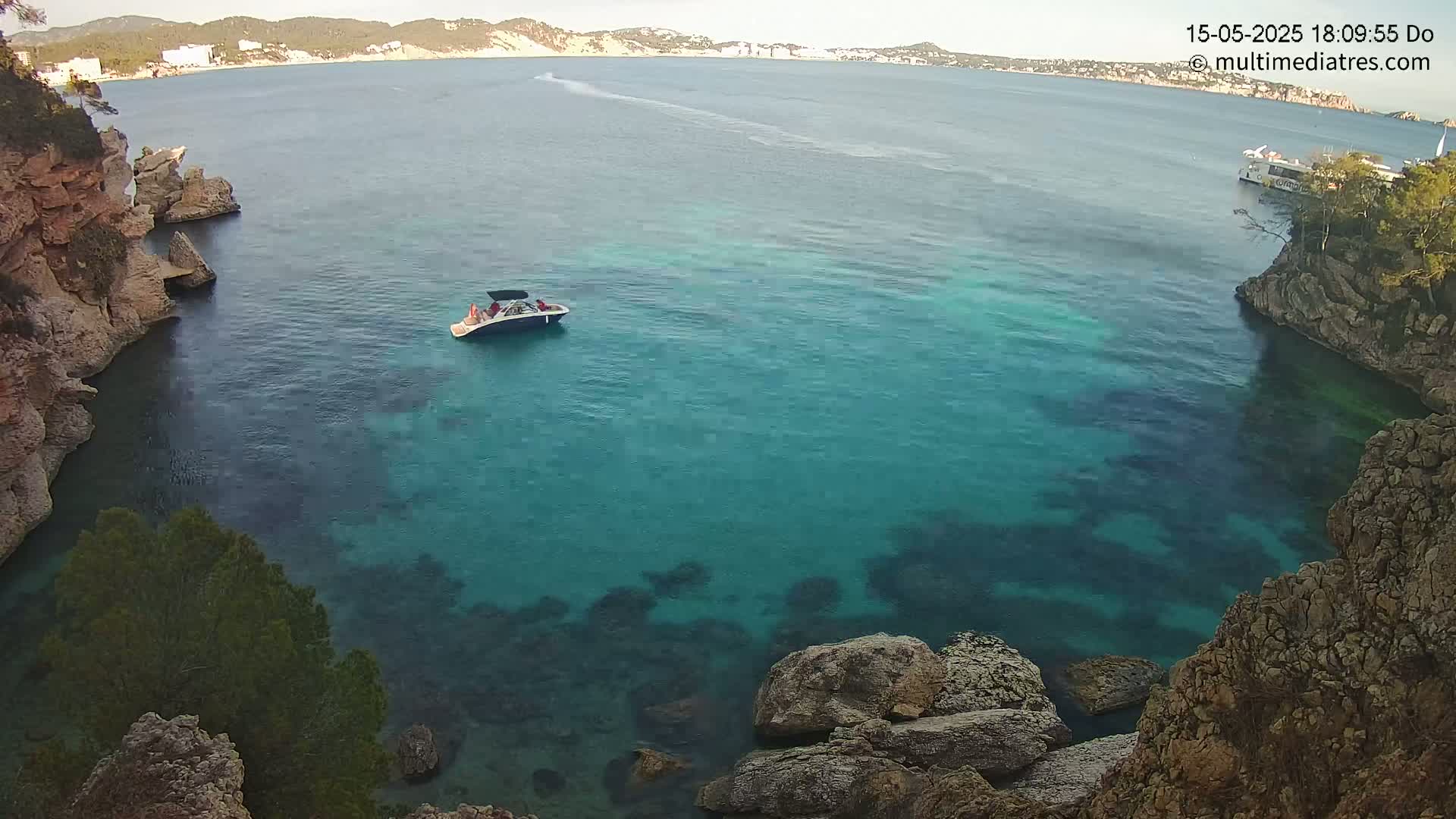 A small boat with people on it sits in a calm, clear turquoise cove surrounded by rocky cliffs under a sunny sky.