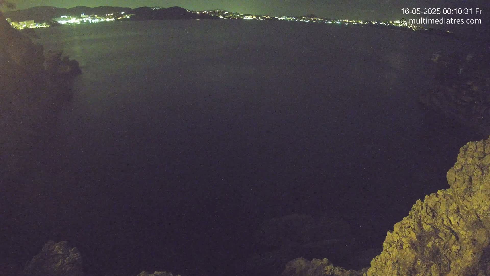 A nighttime view of a dark body of water with a distant, lit cityscape visible across the water, partially obscured by darkness and a rocky foreground.