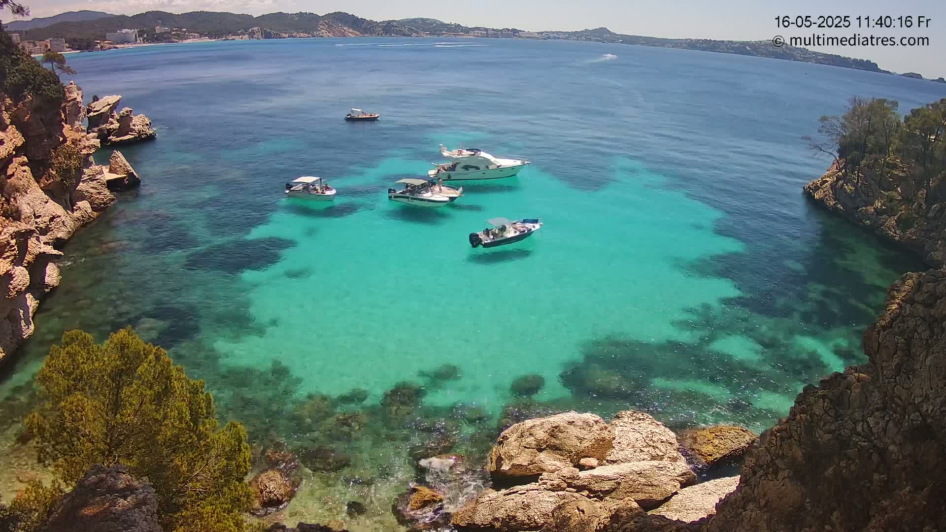 Several boats are anchored in a clear, turquoise cove surrounded by rocky, vegetated shores on a sunny day.