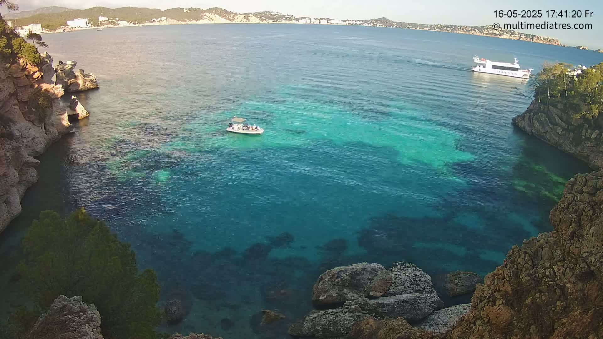 A tranquil cove with clear, turquoise water is nestled between rocky cliffs, with a small boat and a larger vessel visible in the distance under a sunny sky.