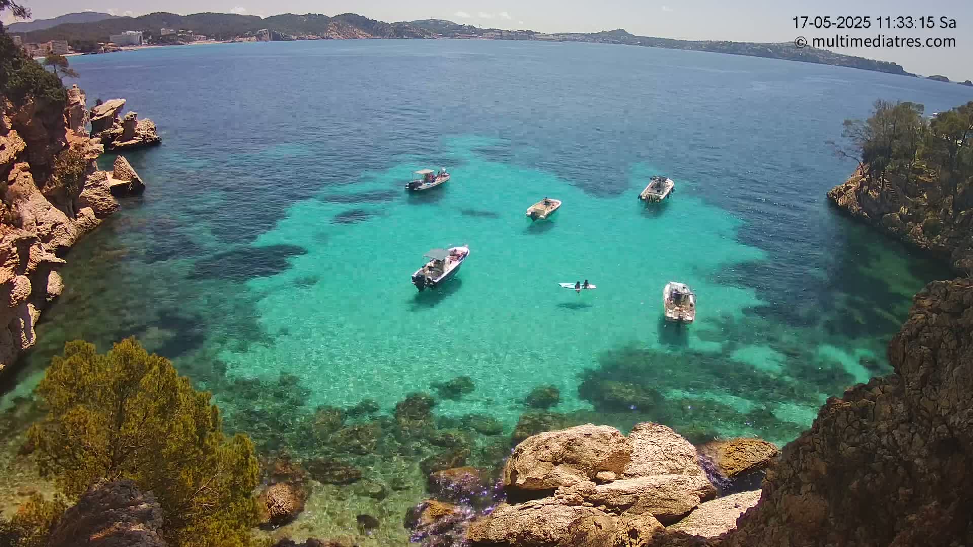 A secluded cove with crystal-clear turquoise water, several small boats, and rocky, vegetated shores under sunny skies.
